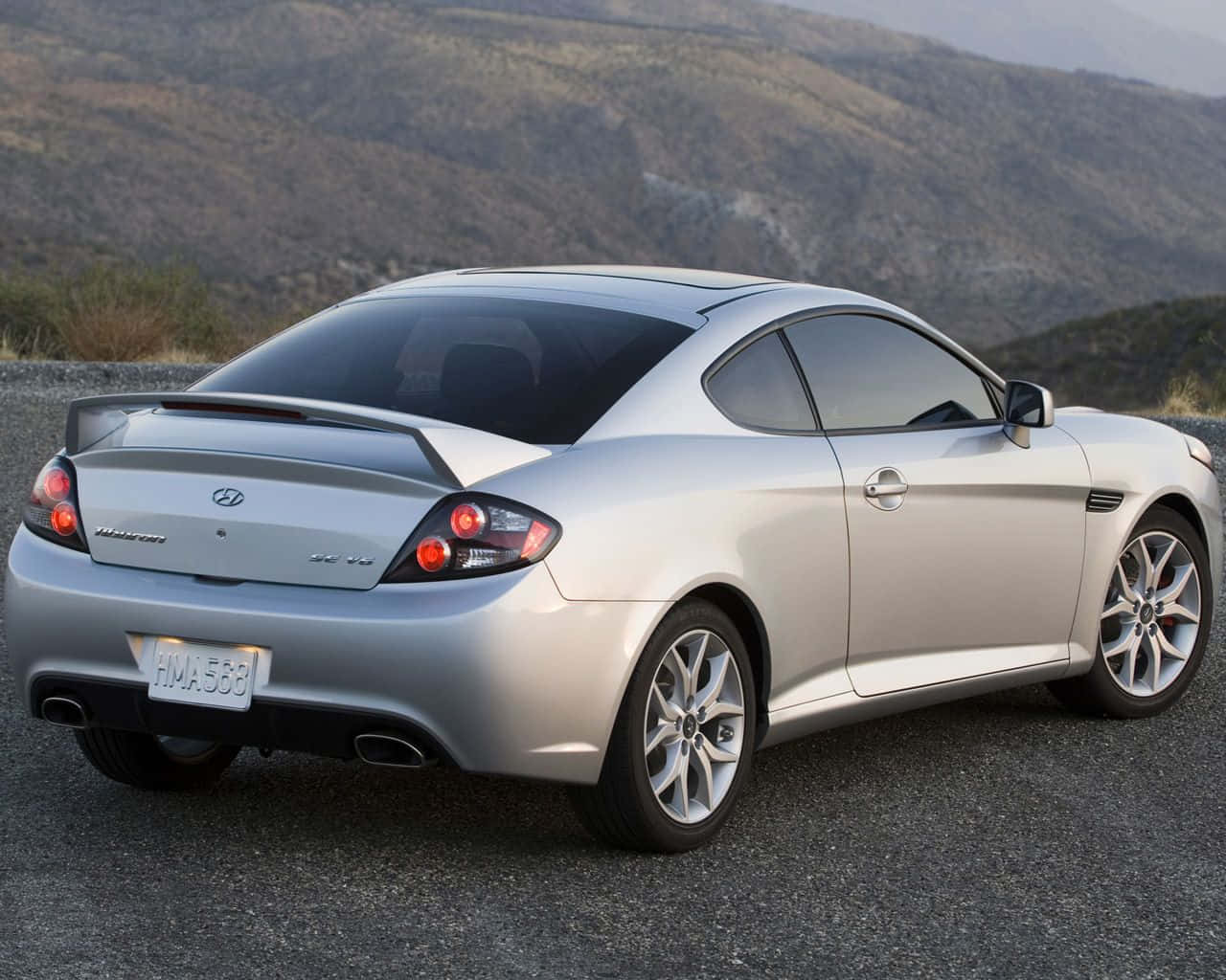 Hyundai Tiburon 桌布 壁紙 — 一輛銀色現代Genesis Coupe停泊在碎石路上，背景是崎嶇的山丘，在柔和日光下。