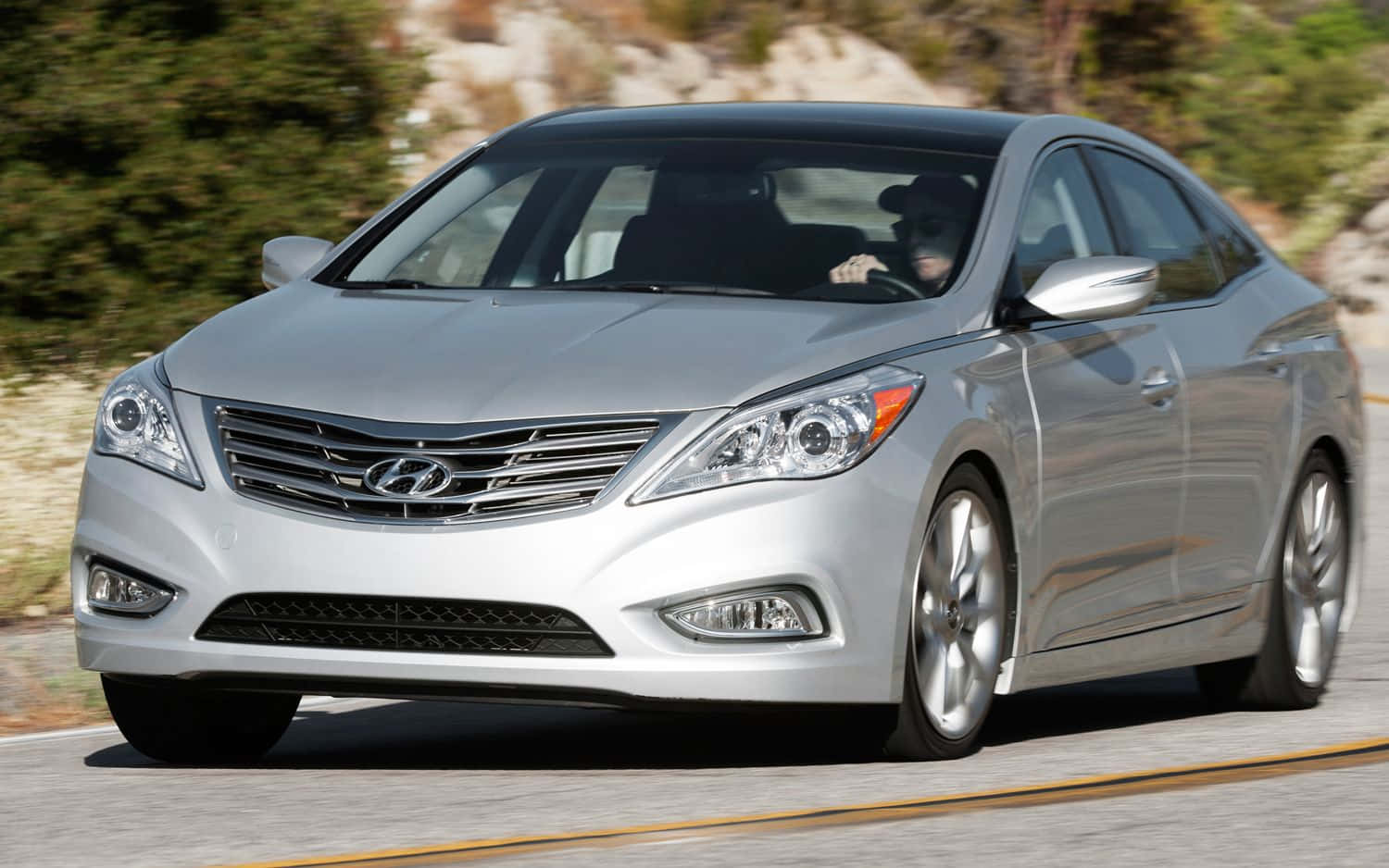 Hyundai Azera 桌布 壁紙 — 一輛銀色現代房車在陽光明媚的蜿蜒道路上行駛，背景是模糊的綠植和岩石地形。