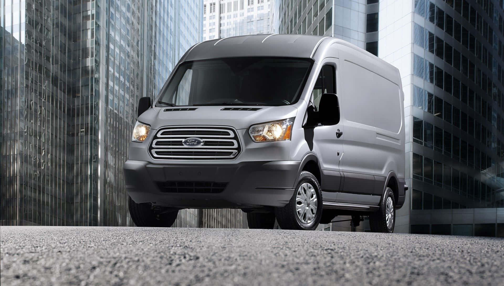 Ford Transit 桌布 壁紙 — 一輛銀色福特 Transit 客貨車停泊在現代城市中高聳玻璃摩天大樓之間的瀝青路面上。