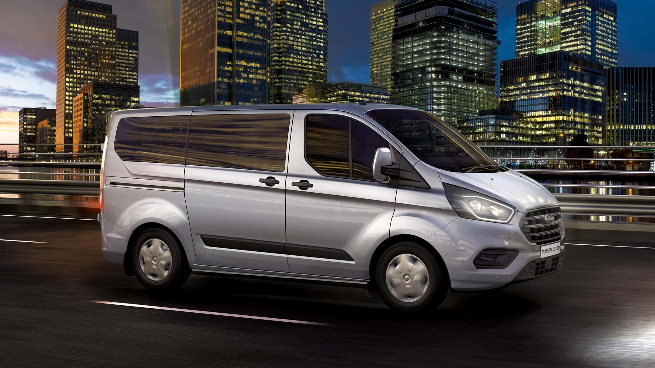 Ford Transit 桌布 壁紙 — 一輛銀色福特Transit客貨車在黃昏時分於城市高速公路上飛馳，背景是燈火通明的摩天大樓。