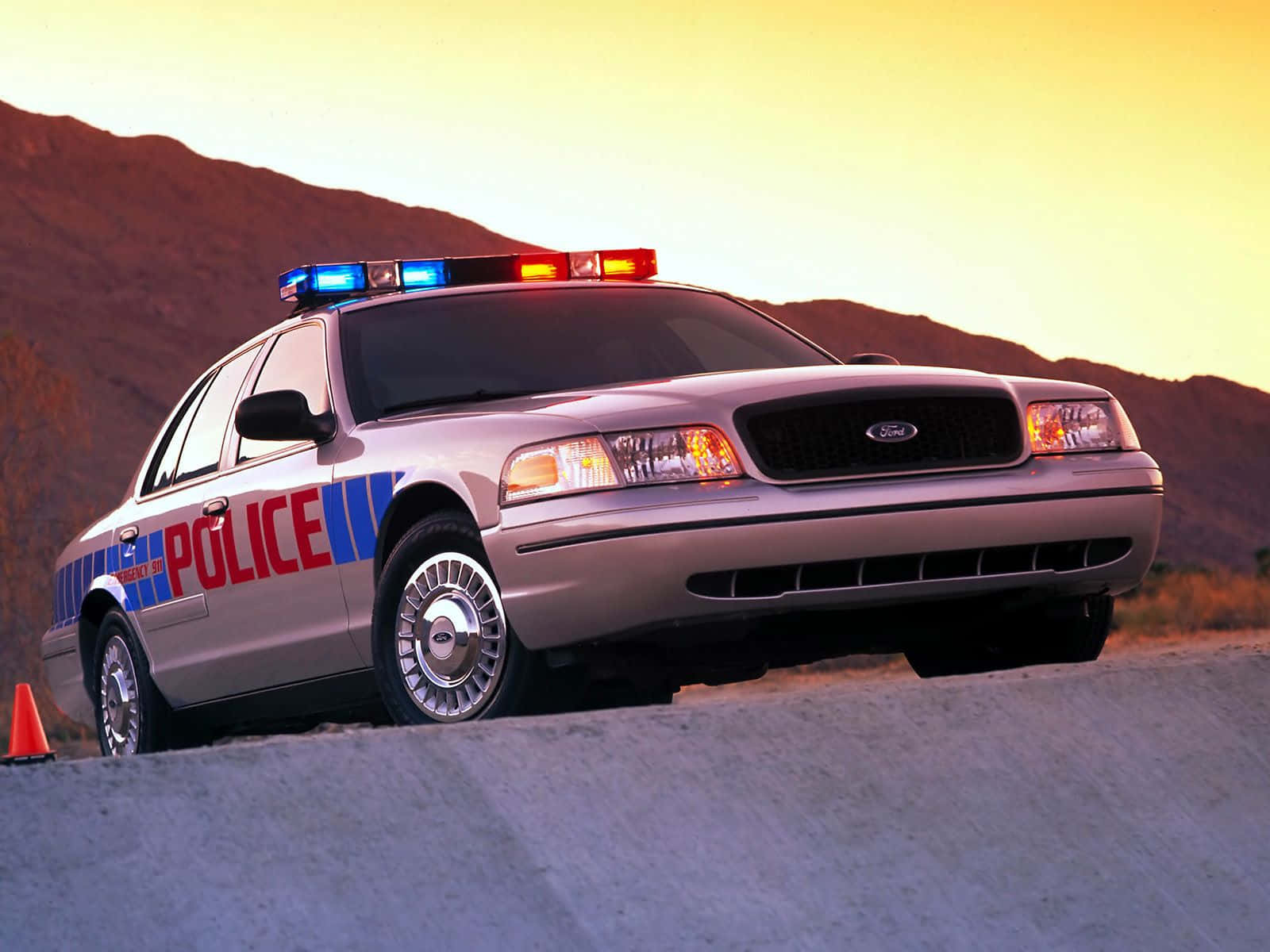 Ford Crown Victoria 桌布 壁紙 — 一輛銀色警車閃著警燈，在日落時分停泊在路邊，背景是山脈。