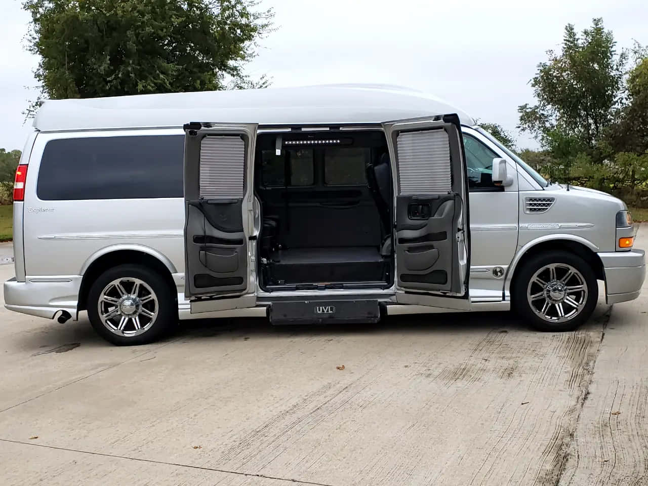 Gmc Savana 桌布 壁紙 — 一輛銀色豪華客貨車打開側門，露出黑色皮革內飾，停泊在樹木環繞的混凝土車道上。
