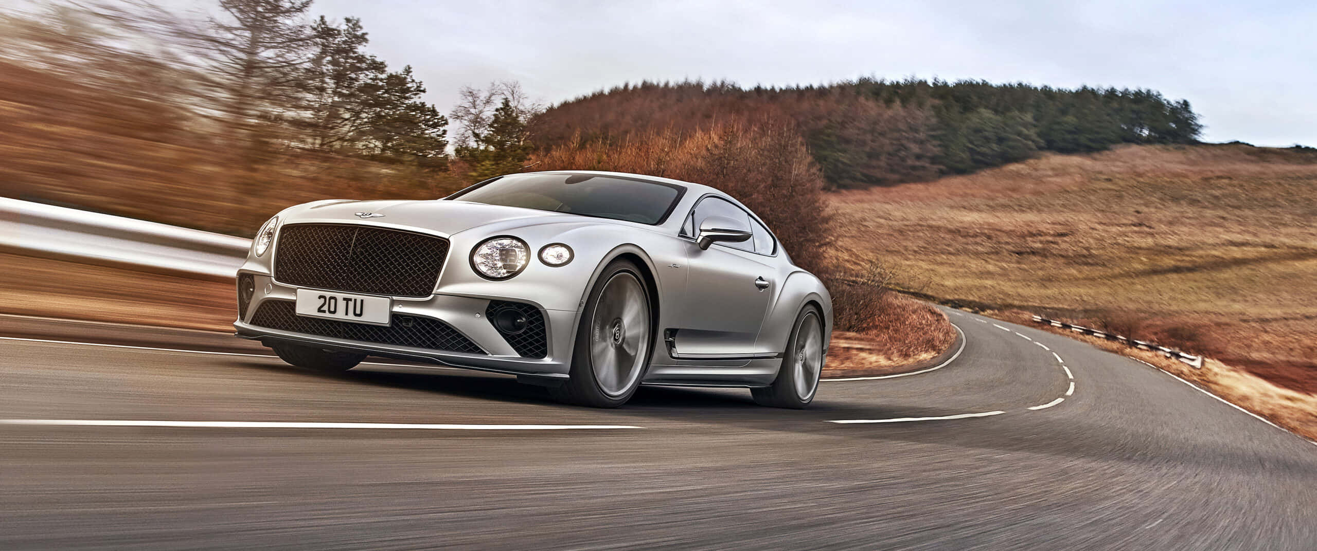 Bentley Continental Gt 桌布 壁紙 — 一輛銀色賓利 Continental GT 在陰天之下，沿著蜿蜒的鄉村道路穿過秋日山丘疾馳。
