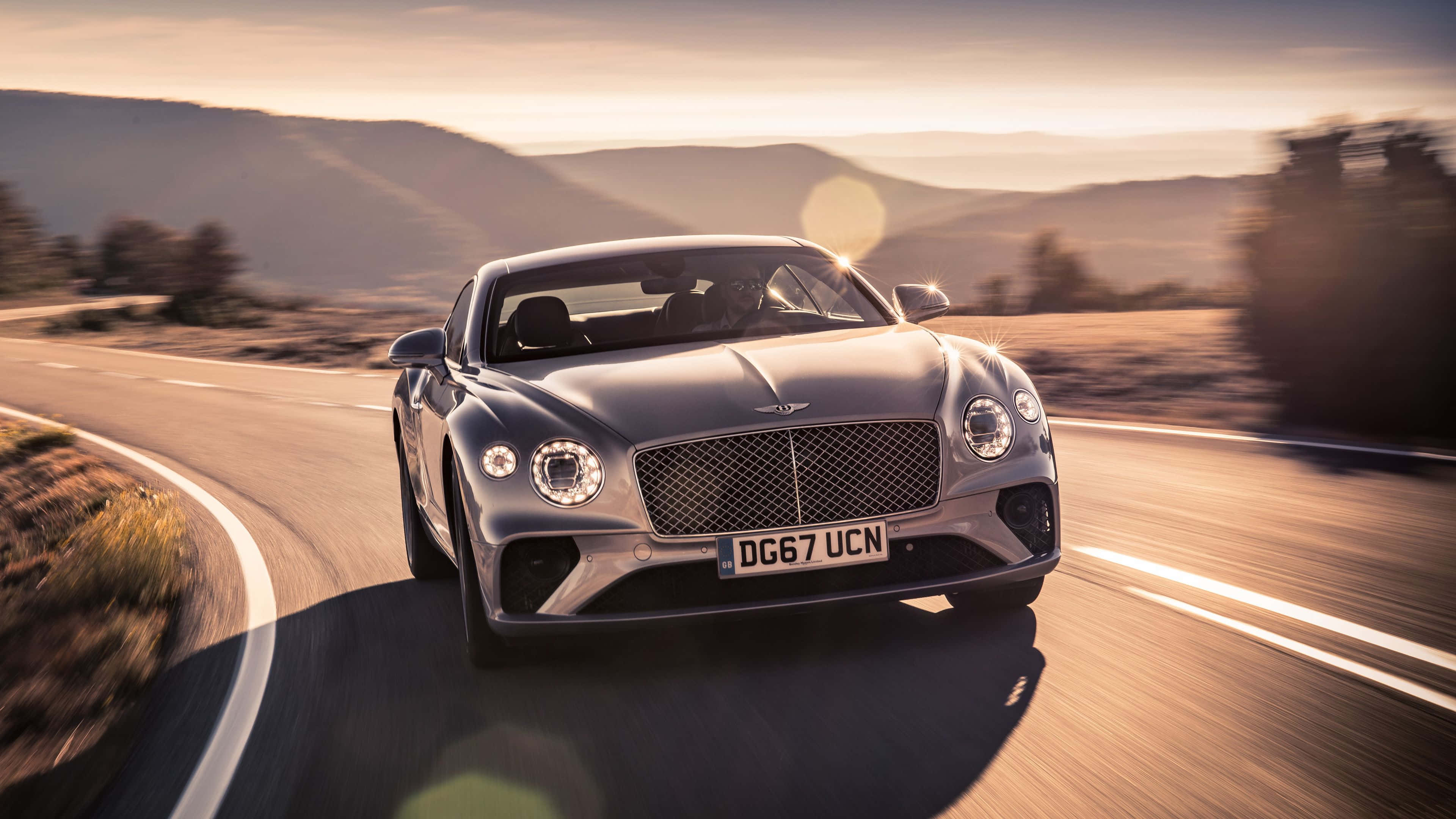 Bentley Continental Gt 桌布 壁紙 — 一輛銀色賓利 Continental GT 在日落時分沿著蜿蜒的山路疾馳，帶有模糊的動感和溫暖的金色光芒。