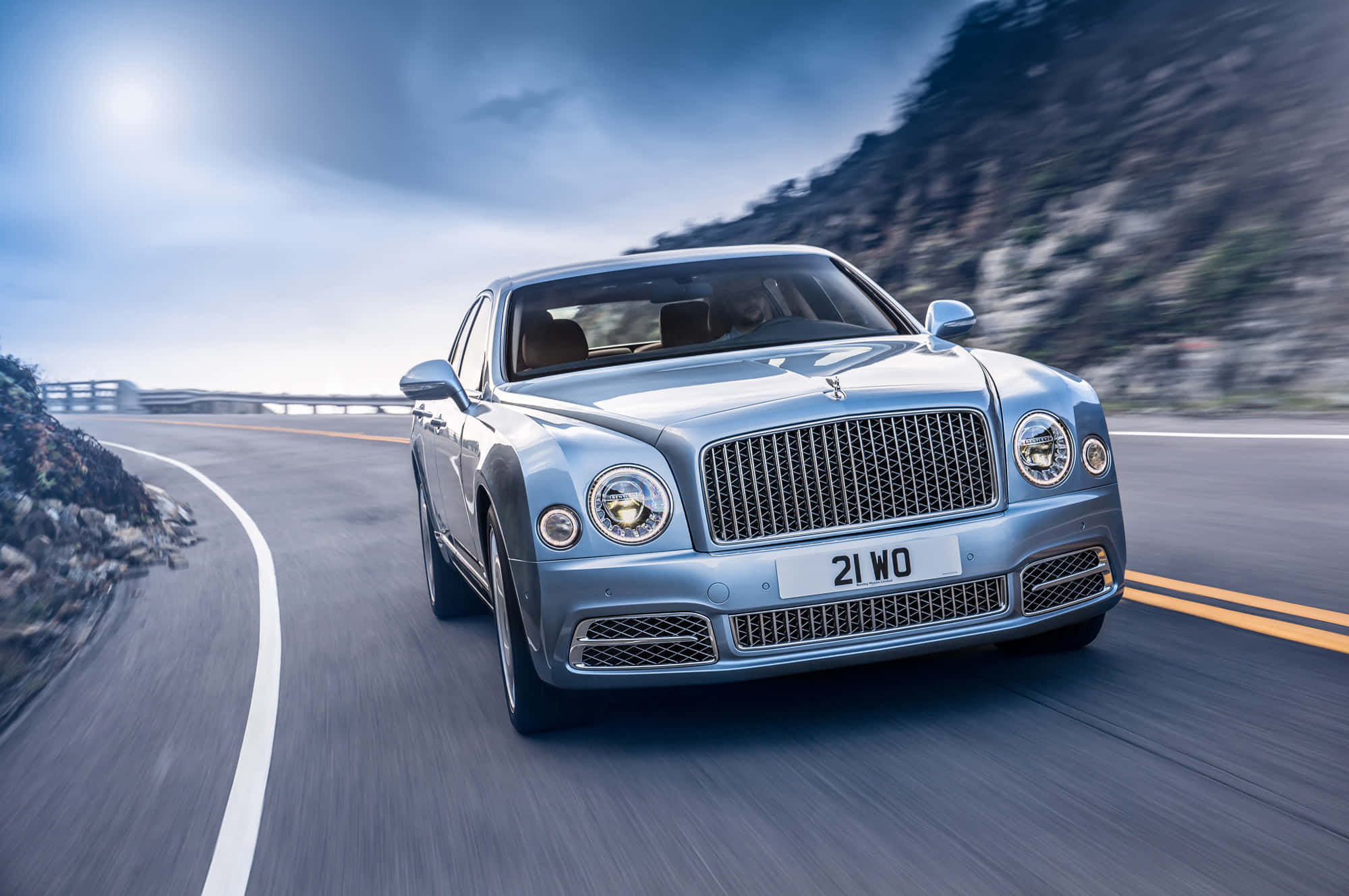 Bentley Mulsanne 桌布 壁紙 — 一輛銀色賓利慕尚在明亮的天空下，沿著蜿蜒的海岸道路疾馳，帶有模糊的動態和岩石峭壁。