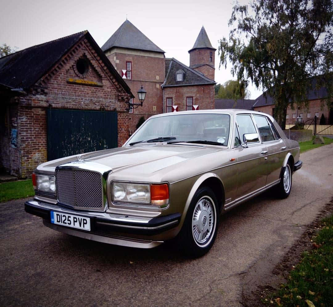 Bentley Eight 桌布 壁紙 — 一輛銀色賓利房車停泊在碎石車道上，前方是一座帶有塔樓和樹木的歷史磚砌莊園。