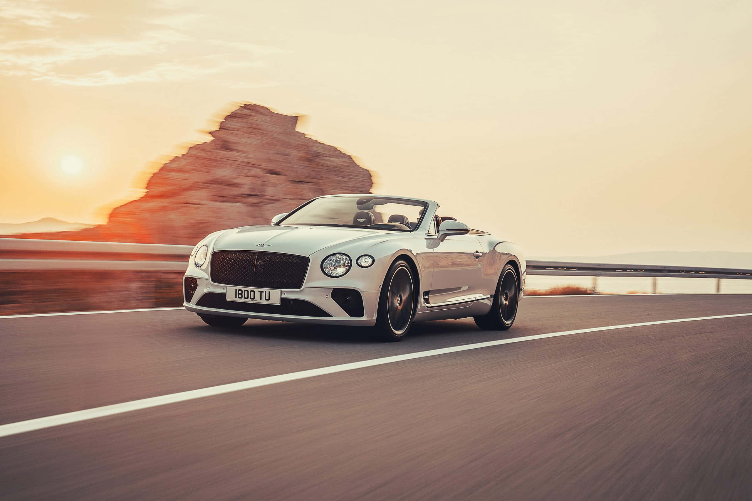 Bentley Continental Gt Convertible 桌布 壁紙 — 一輛銀色賓利開篷跑車在日落時分沿沙漠高速公路飛馳，背景有模糊的岩石形成。