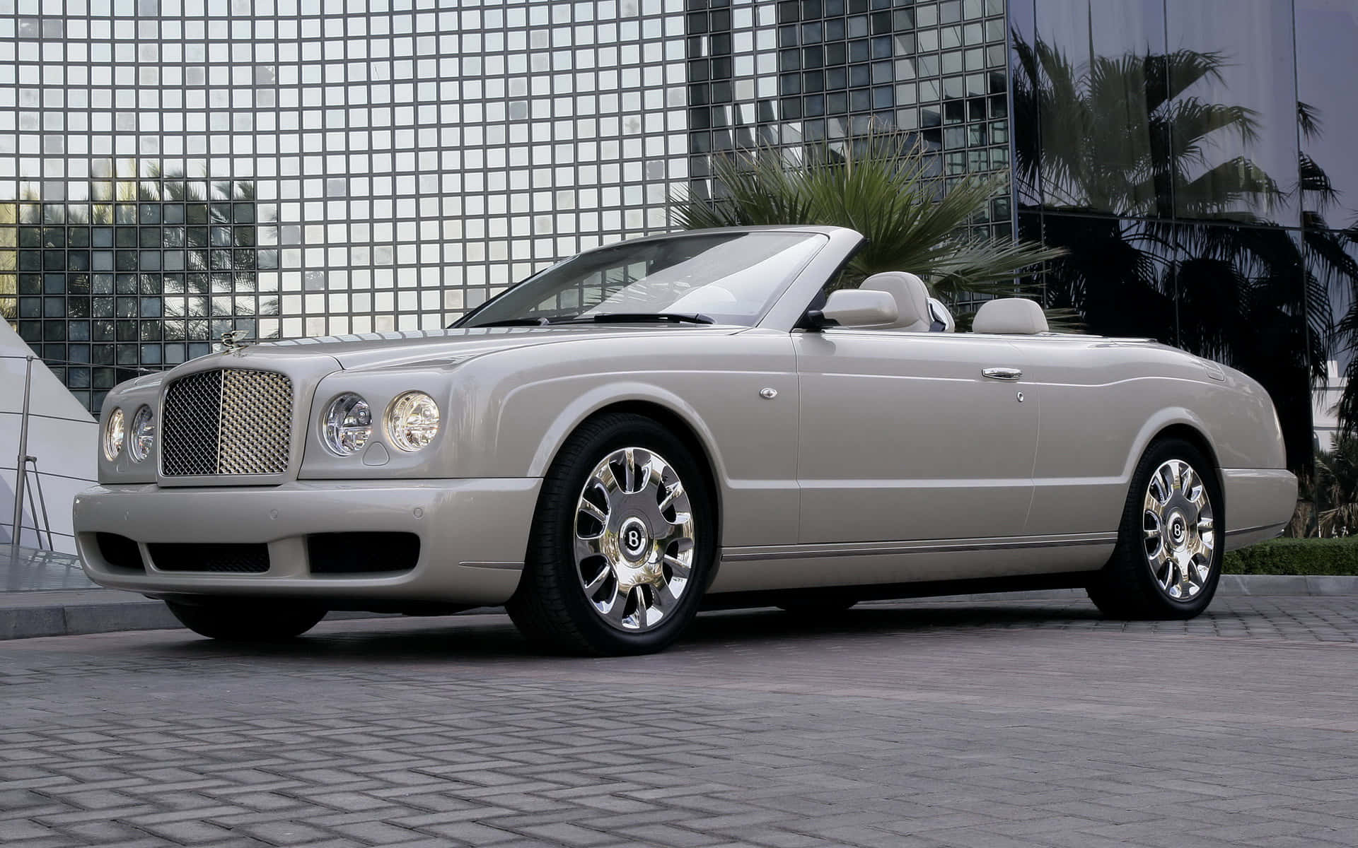 Bentley Azure 桌布 壁紙 — 一輛銀色賓利開篷車停泊在鋪砌的廣場上，前方是一座玻璃摩天大樓，棕櫚樹倒映在窗戶中。