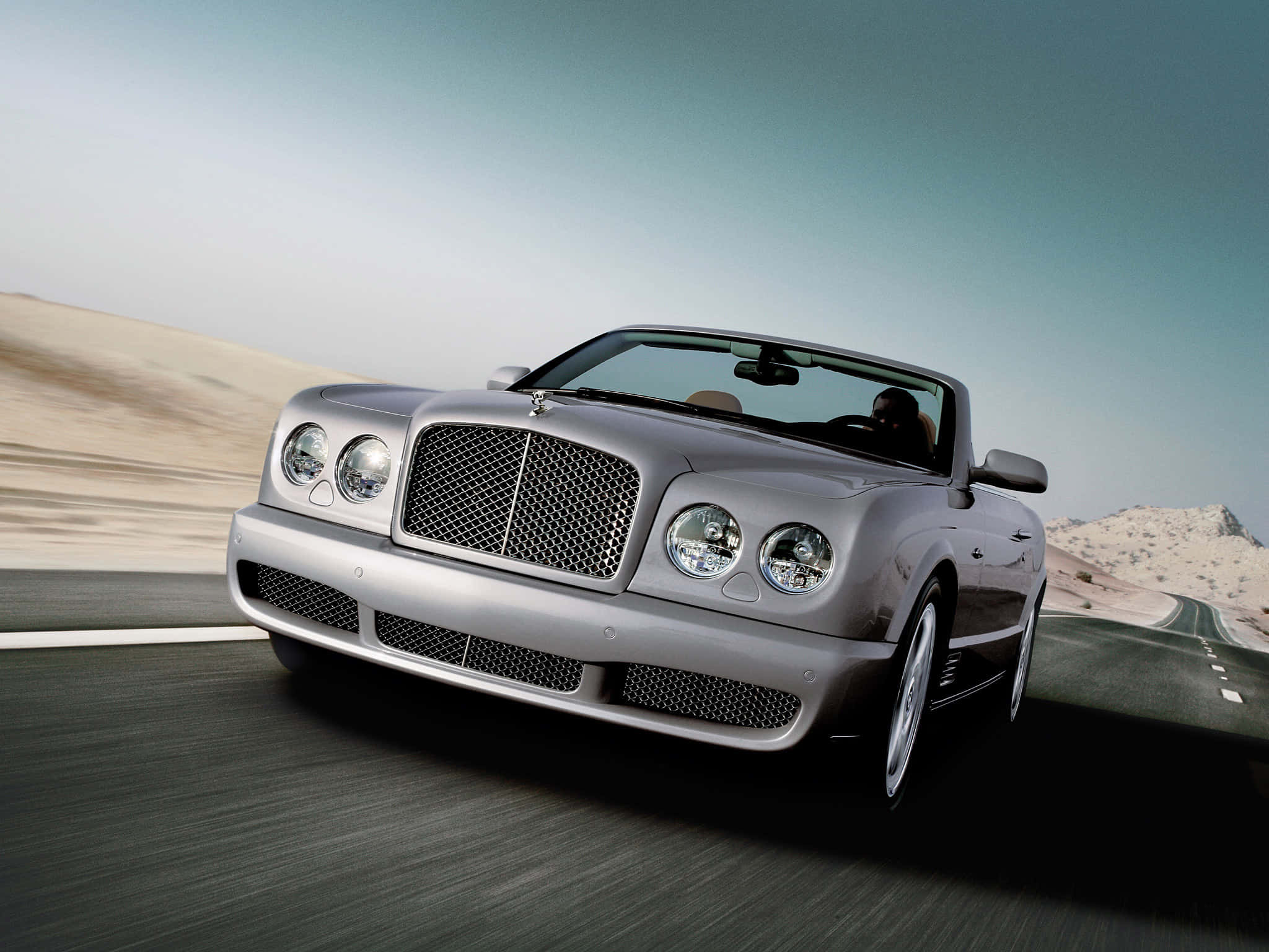 Bentley Azure 桌布 壁紙 — 一輛銀色賓利開篷車在晴朗天空下，沿沙漠公路高速行駛，呈現模糊動感及岩石地形。