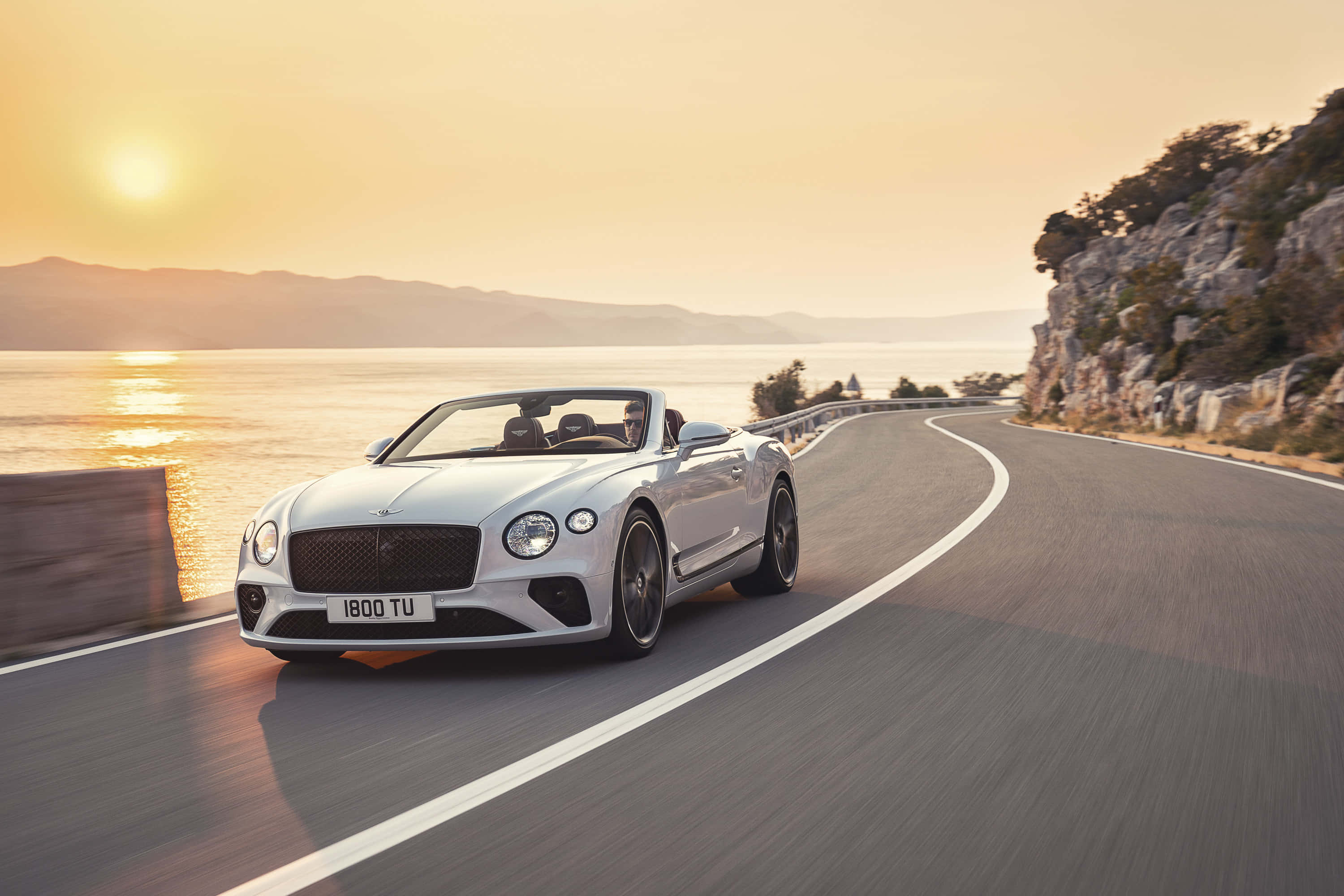 Bentley Continental Gt Convertible 桌布 壁紙 — 一輛銀色賓利開篷車沿著海岸公路在日落時分巡航，金色光芒在水面上反射。
