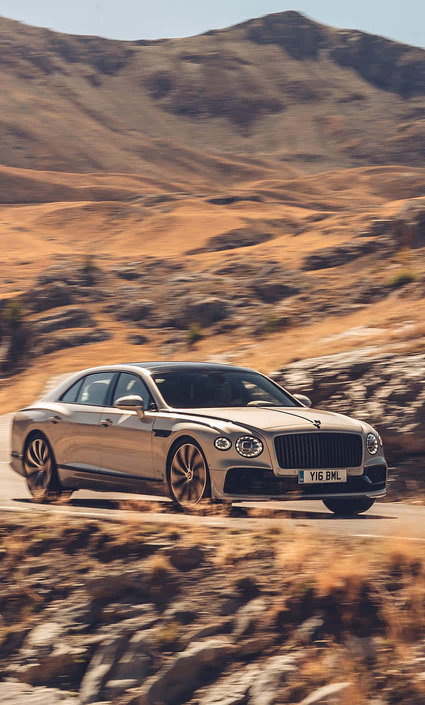 Bentley Flying Spur 桌布 壁紙 — 一輛銀色賓利飛馳在蜿蜒的山路上，穿越乾燥的金色山丘，在明亮的日光下行駛。