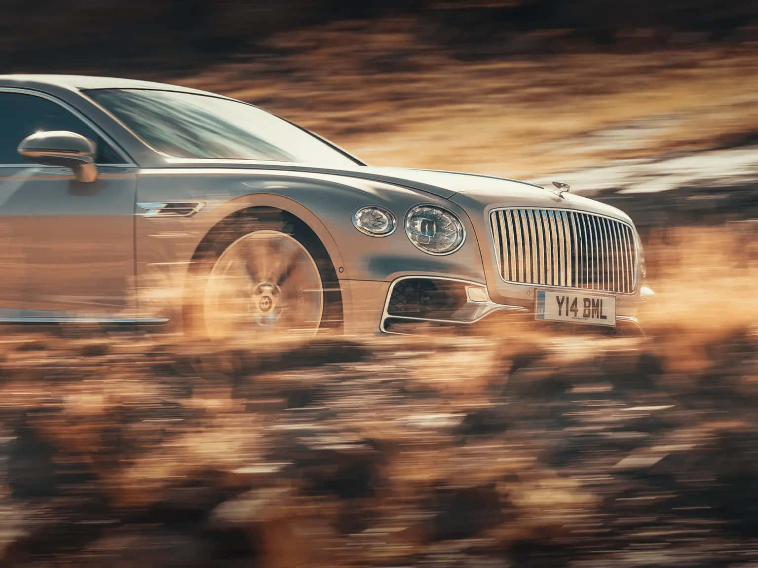 Bentley Flying Spur 桌布 壁紙 — 一輛銀色賓利飛馳在金色沙漠景觀中高速行駛，動態模糊強調了速度。