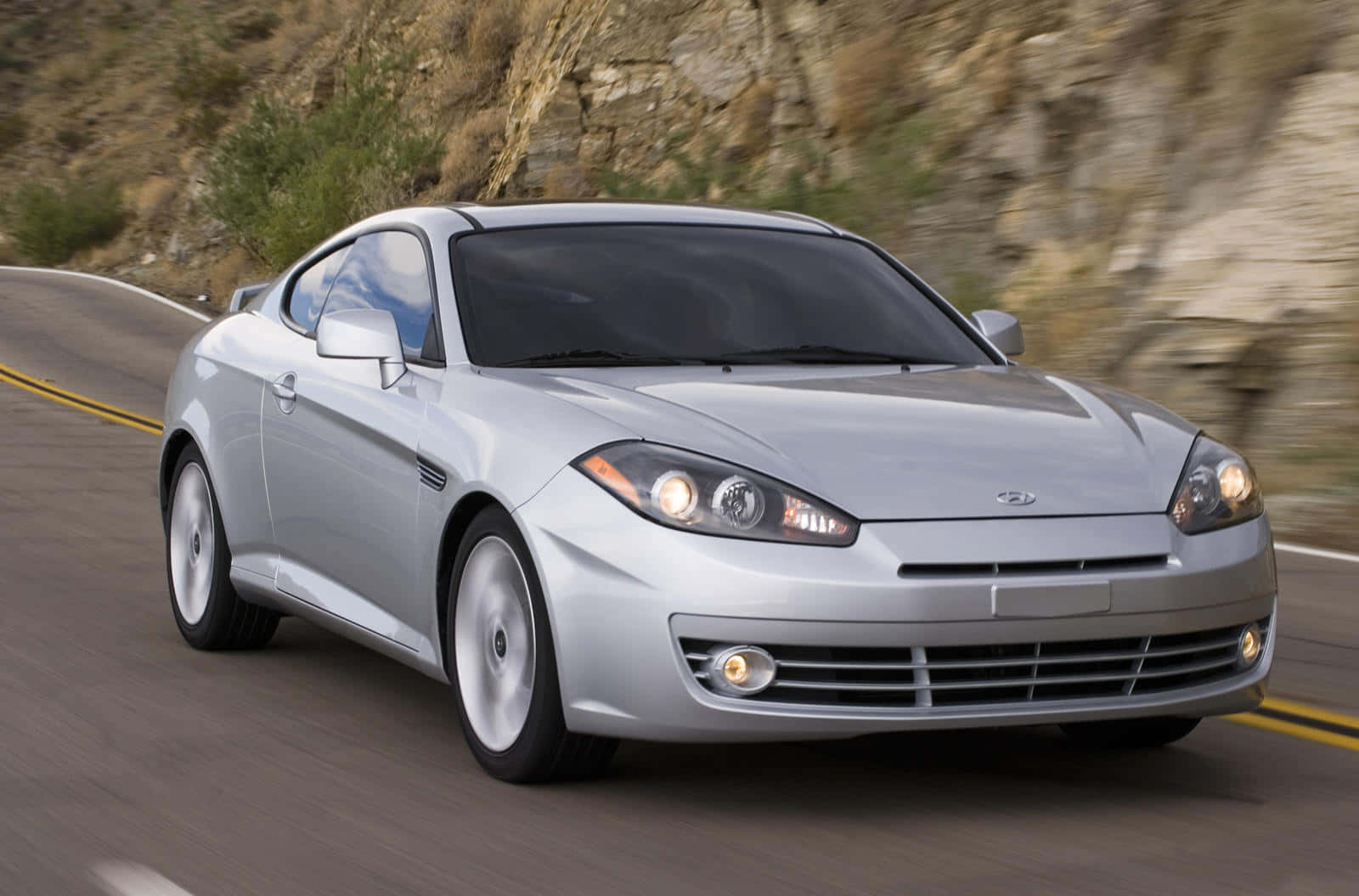 Hyundai Tiburon 桌布 壁紙 — 一輛銀色跑車沿著蜿蜒的山路疾馳，背景是模糊的岩石峭壁。