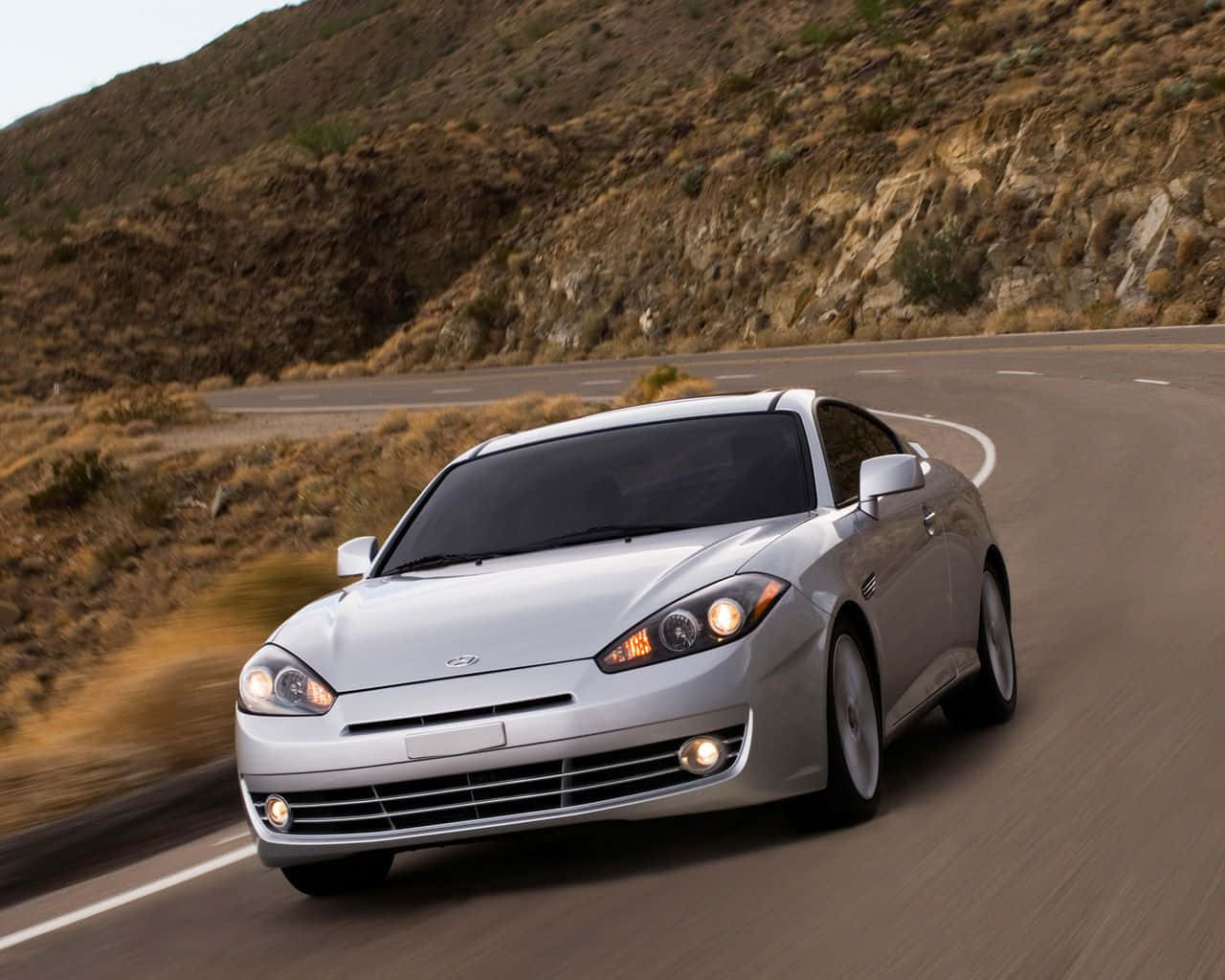 Hyundai Tiburon 桌布 壁紙 — 一輛銀色跑車沿著蜿蜒的山路飛馳，背景是乾燥的灌木叢和岩石地形。