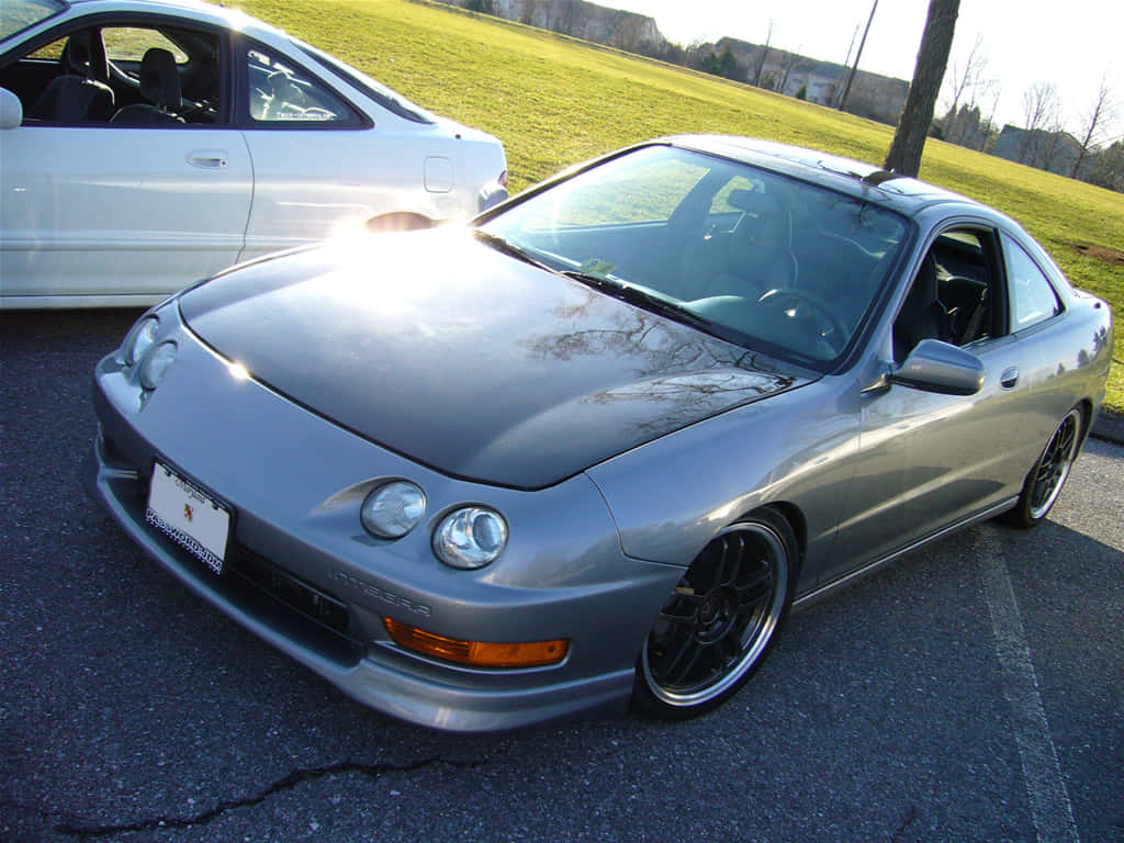Honda Integra 桌布 壁紙 — 一輛銀色跑車配備定制輪圈，在陽光下閃閃發光，停在一輛白色轎車旁邊，位於裂開的瀝青停車場上。