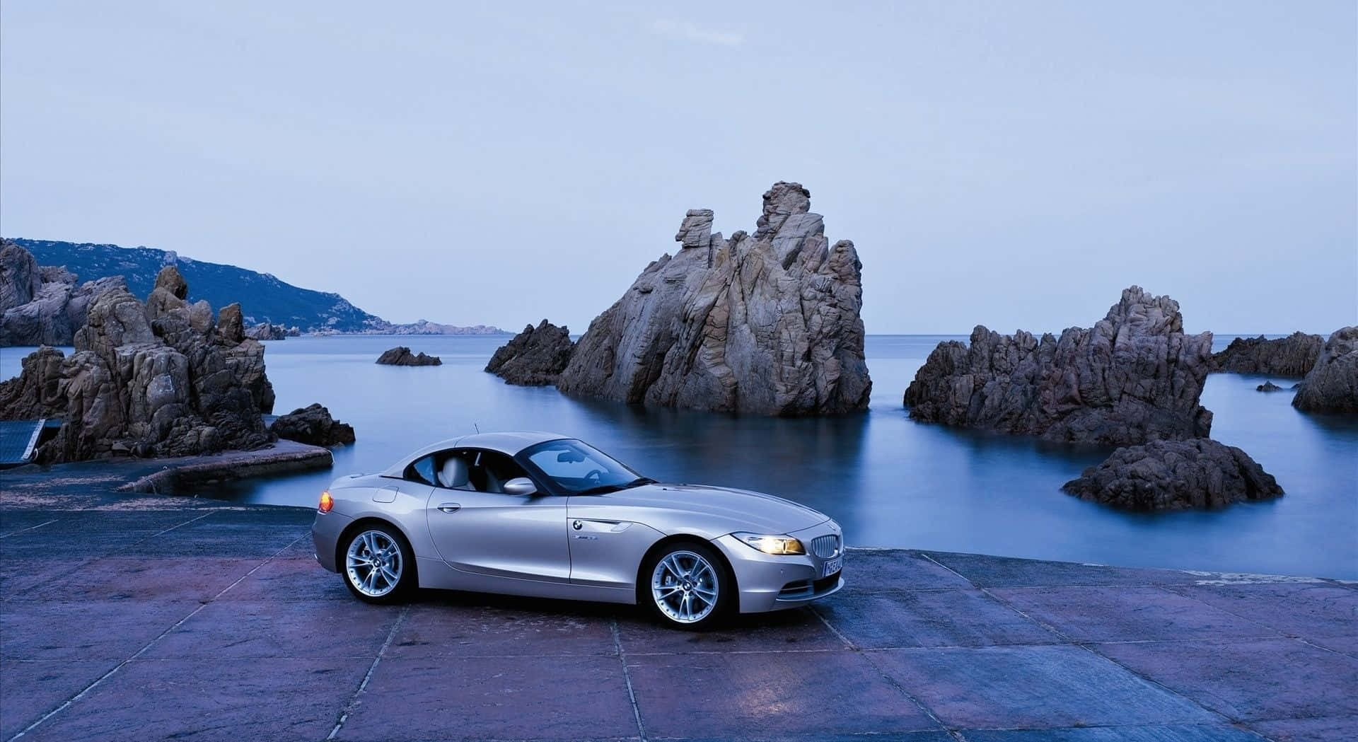 Bmw Z4 桌布 壁紙 — 一輛銀色開篷跑車停泊在石碼頭旁，旁邊是平靜的海水，黃昏時分有戲劇性的岩石 formations。