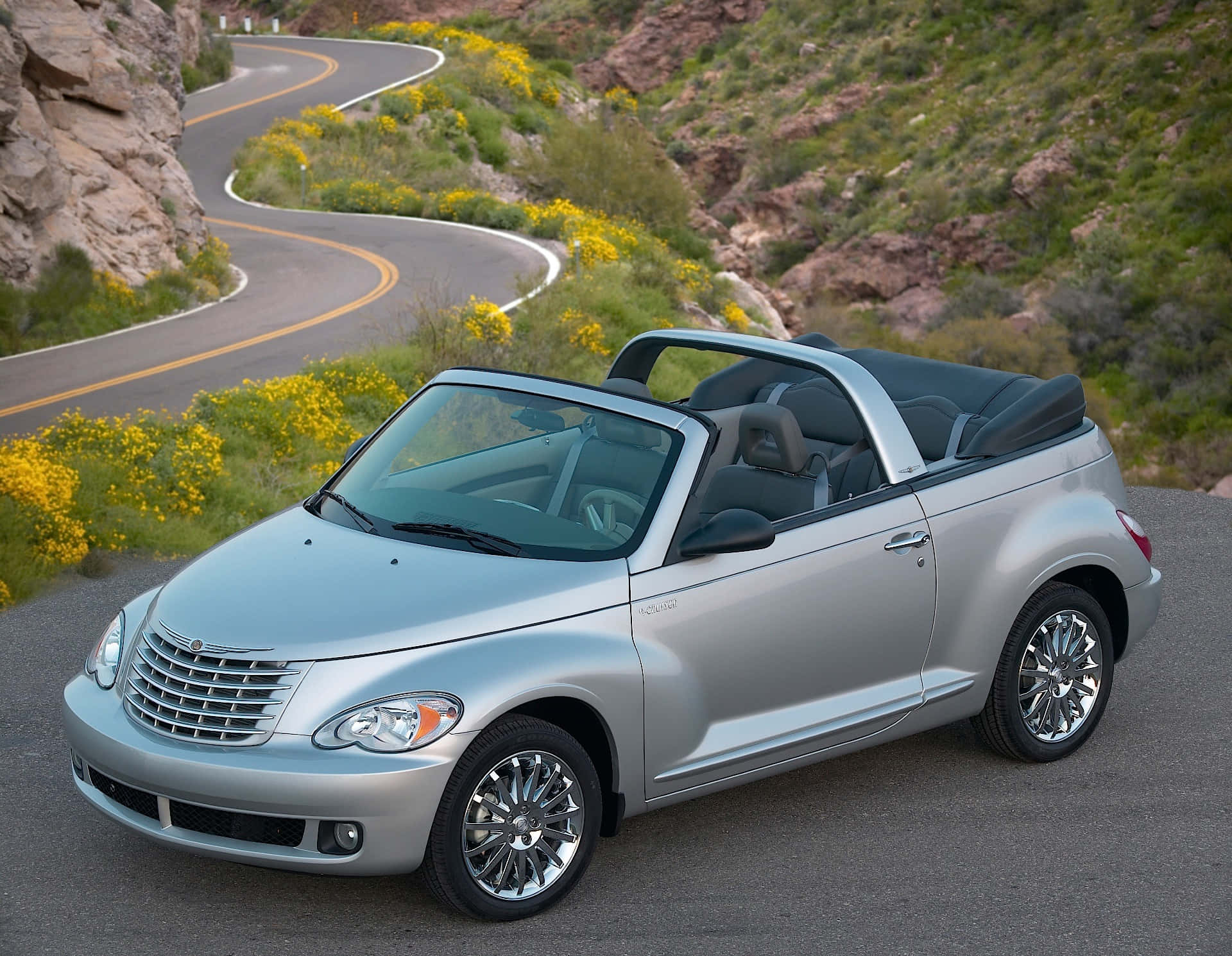 Chrysler Pt Cruiser 桌布 壁紙 — 一輛銀色開篷跑車停泊在蜿蜒的山路上，周圍是黃色野花和岩石地形。