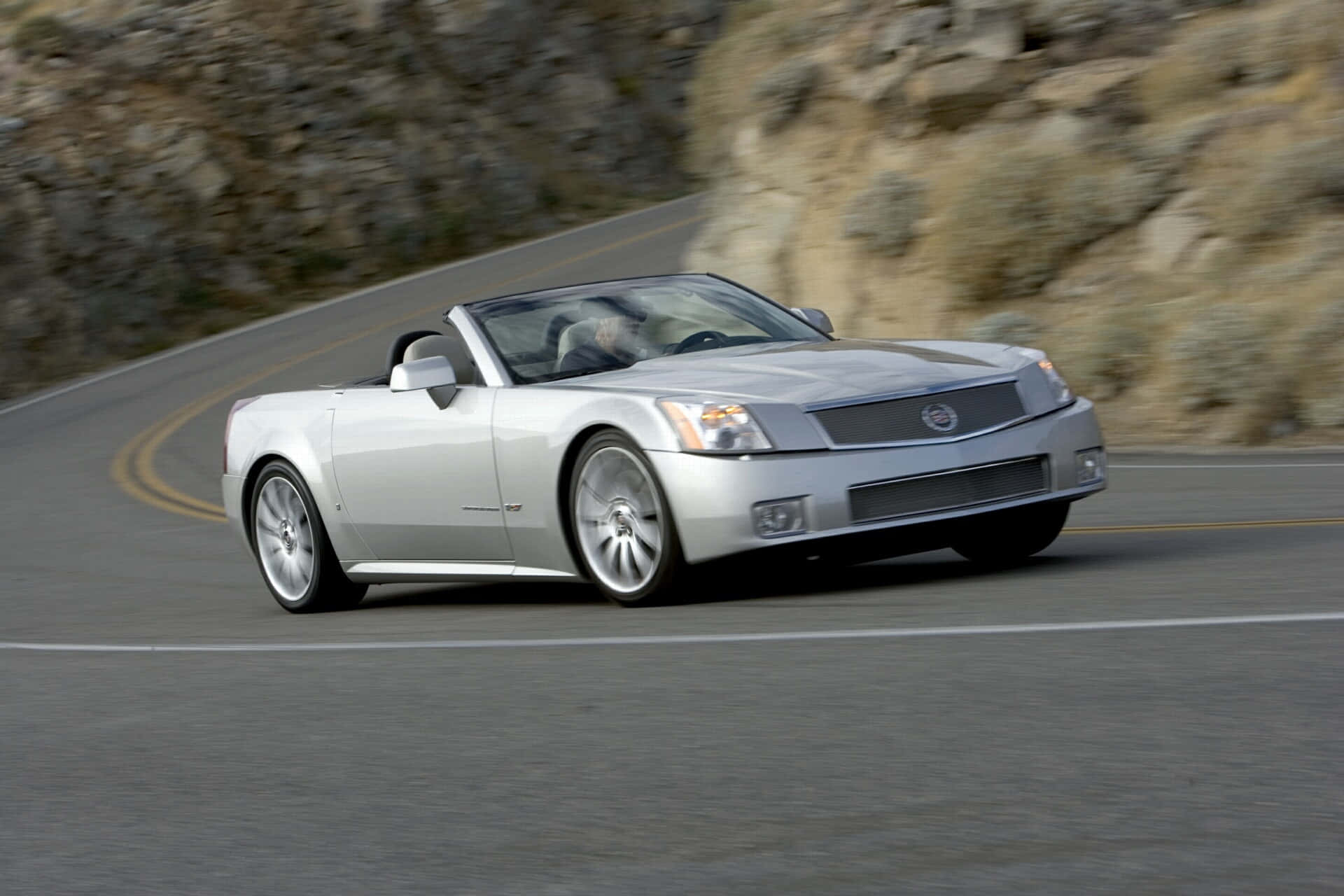 Cadillac Xlr 桌布 壁紙 — 一輛銀色開篷跑車沿著蜿蜒的山路飛馳，背景是模糊的岩石懸崖。
