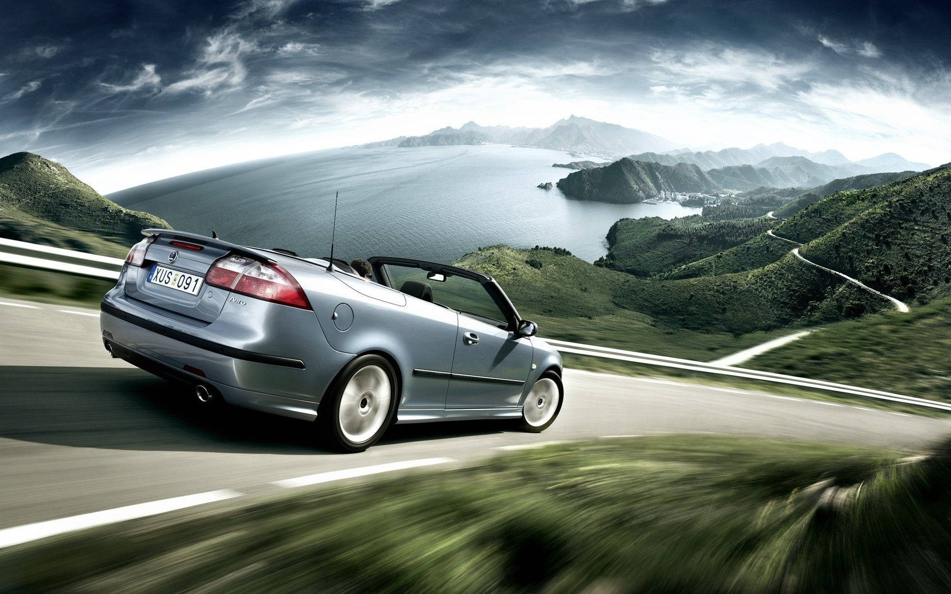 2012 Saab 9-3 Convertible Turbo 壁紙 — 一輛銀色開篷車沿著海岸公路飛馳，兩旁是鬱鬱蔥蔥的綠色山丘和廣闊的藍色大海，天空烏雲密佈，氣氛戲劇化。