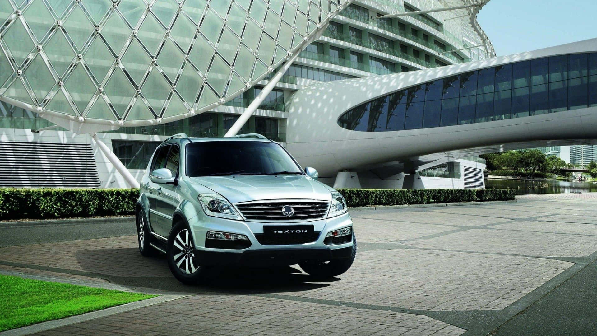 Ssangyong 桌布 壁紙 — 一輛銀色雙龍Tecton SUV停泊在鋪砌的廣場上，前方是一座未來主義風格的玻璃和鋼結構建築，帶有弧形建築設計。