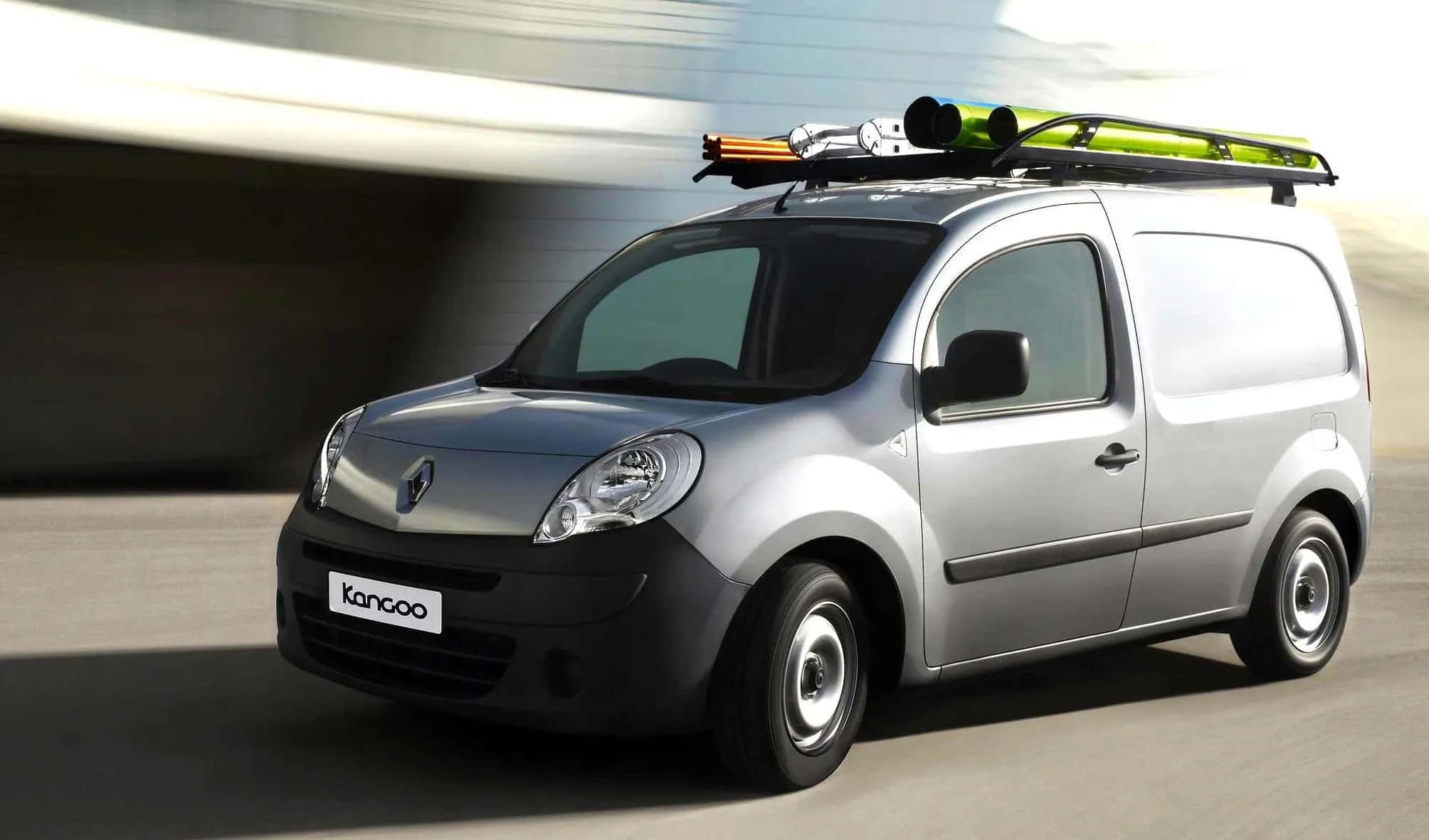 Renault Kangoo 桌布 壁紙 — 一輛銀色雷諾Kangoo客貨車在模糊的道路上行駛，車頂架上綁著綠色和橙色的滑雪板。