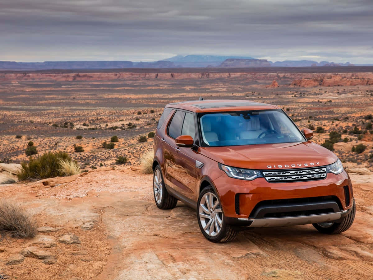 Land Rover Discovery 桌布 壁紙 — 一輛銅色的Land Rover Discovery停泊在岩石沙漠小徑上，遠處有平頂山，天空多雲。