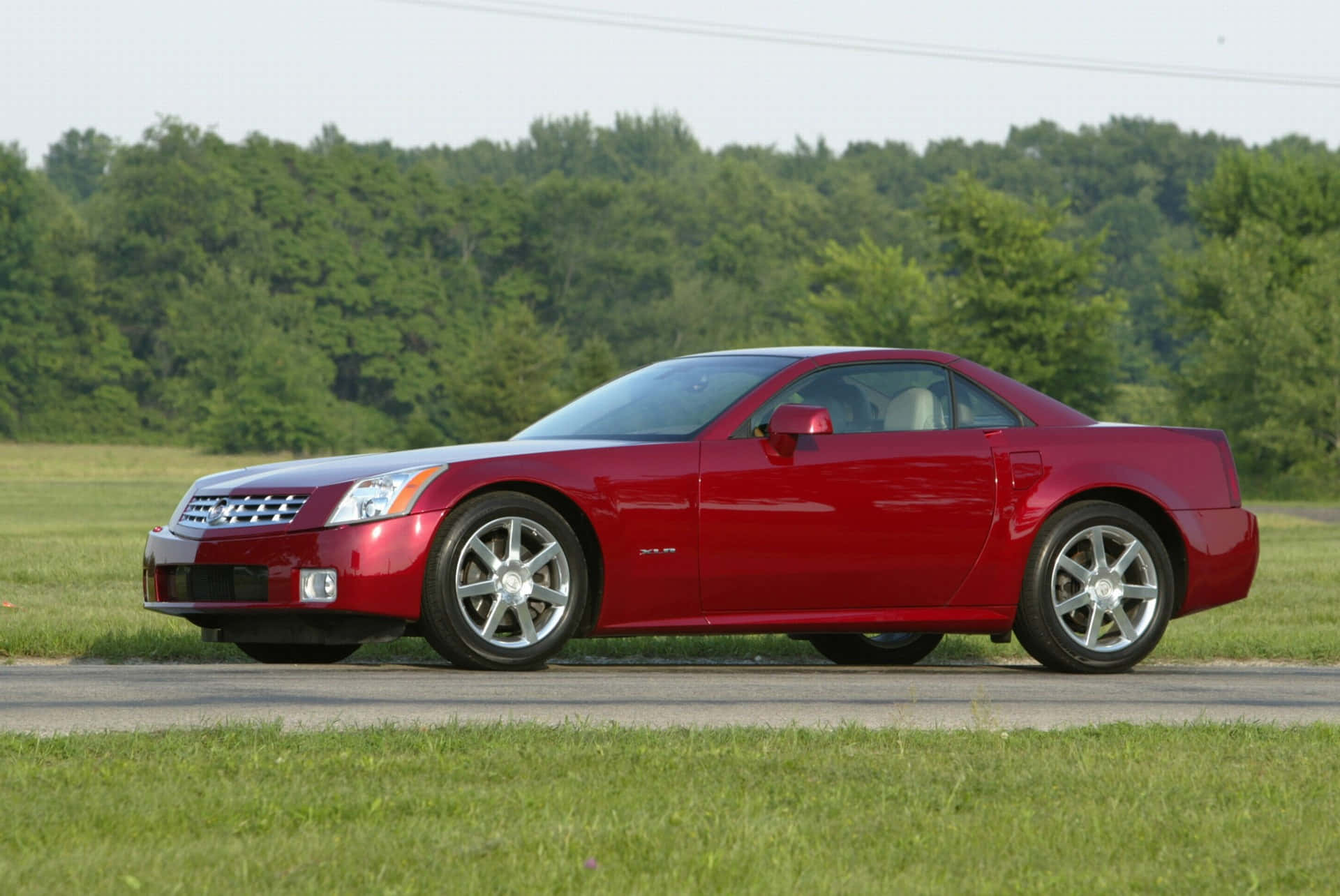 Cadillac Xlr 桌布 壁紙 — 一輛閃亮的紅色 Cadillac XLR 雙門跑車停在路邊，背景是茂密的綠樹和草地。