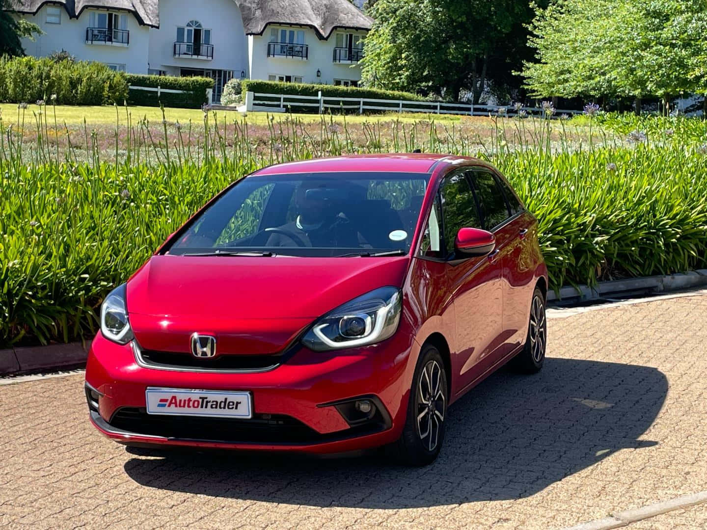 Honda Fit 桌布 壁紙 — 一輛閃亮的紅色本田Jazz停放在鋪砌的車道上，背景是茂密的綠植和一棟白色房屋，在明亮的陽光下。