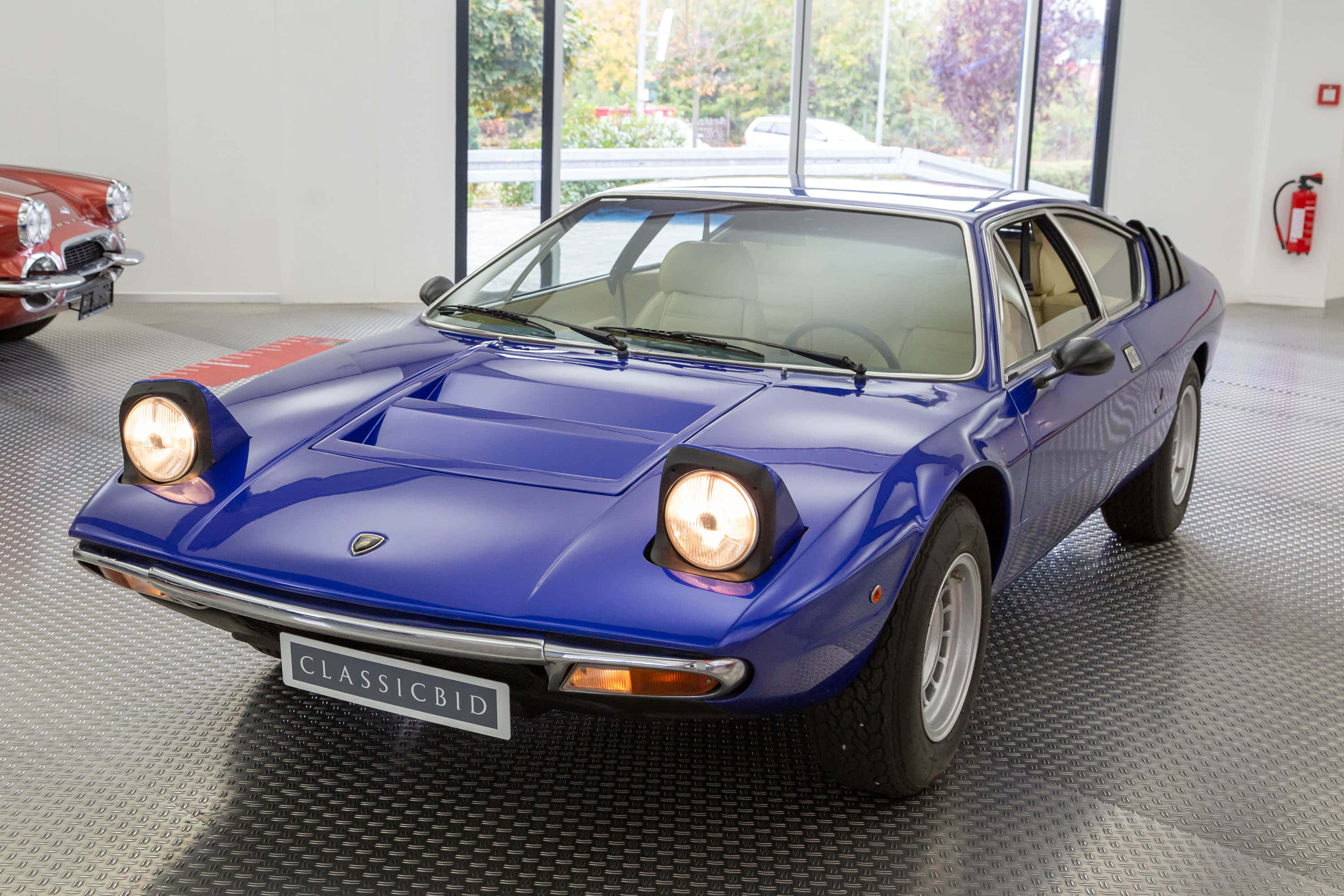 Lamborghini Urraco 桌布 壁紙 — 一輛閃亮的藍色經典林寶堅尼Countach跑車在現代展廳的明亮燈光下閃耀，背景可見一輛紅色汽車。