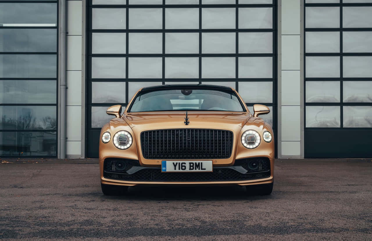 Bentley Flying Spur 桌布 壁紙 — 一輛閃亮的金色賓利 Continental GT 停在一棟帶有大型玻璃門的現代工業建築前。