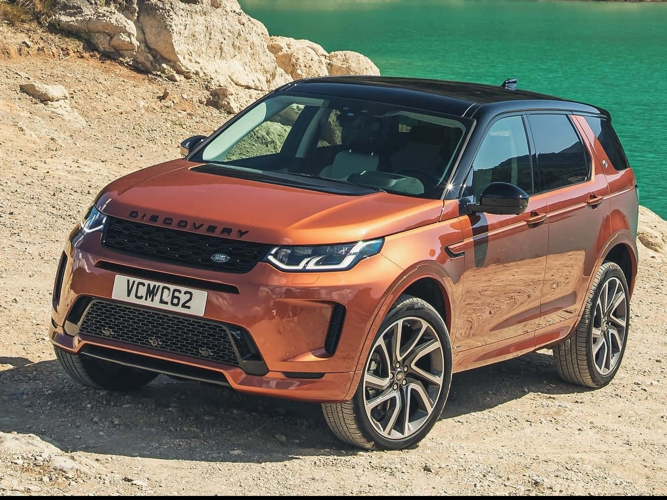 Land Rover Discovery 桌布 壁紙 — 一輛閃亮的銅色 Land Rover Discovery 停泊在岩石地形上，旁邊是充滿活力的綠松石色湖泊，在明亮的日光下。