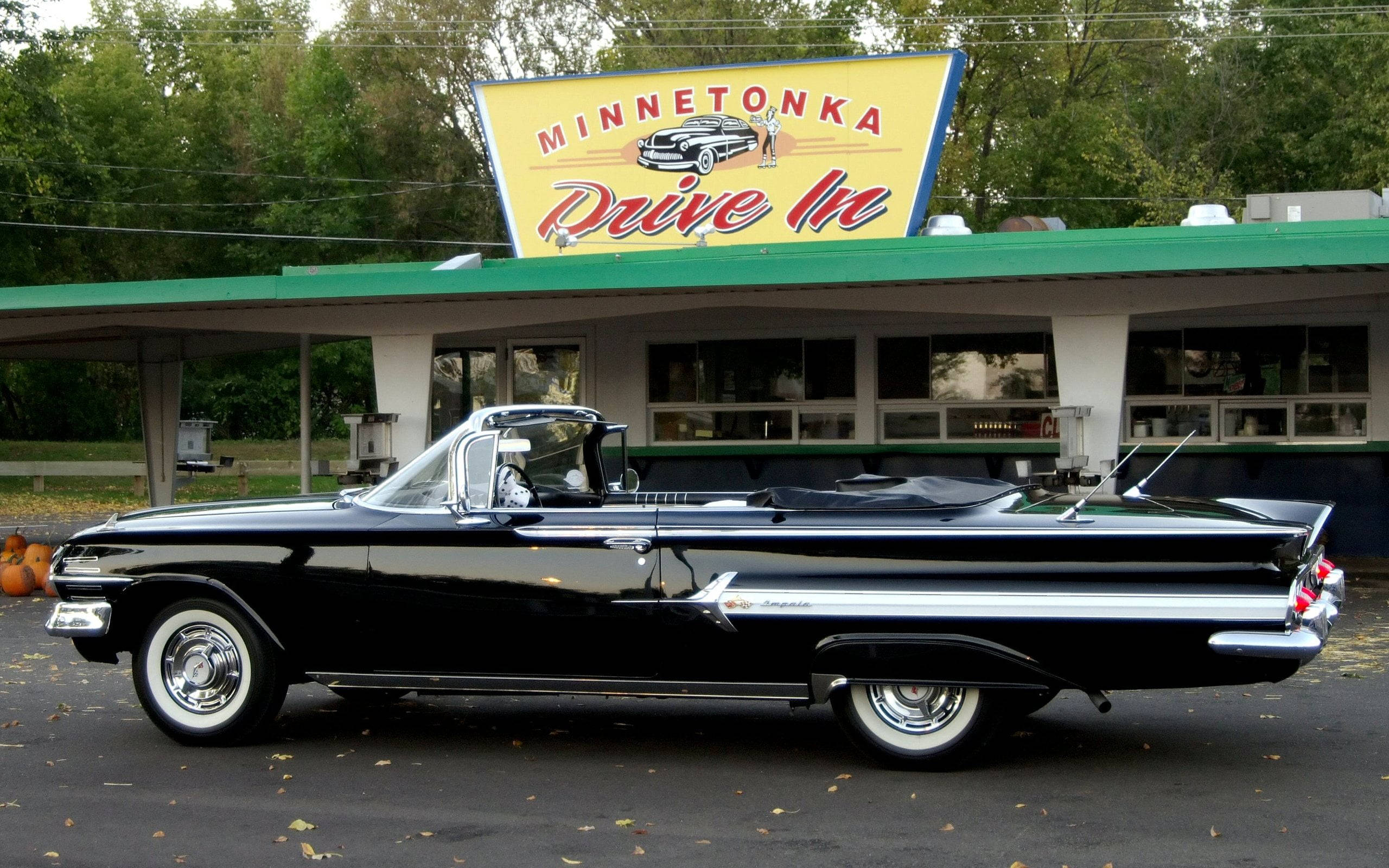 HD 桌布 壁紙 — 一輛閃亮的黑色1950年代開篷車停泊在復古的Minnetonka Drive In前，地上散落著秋天的落葉。