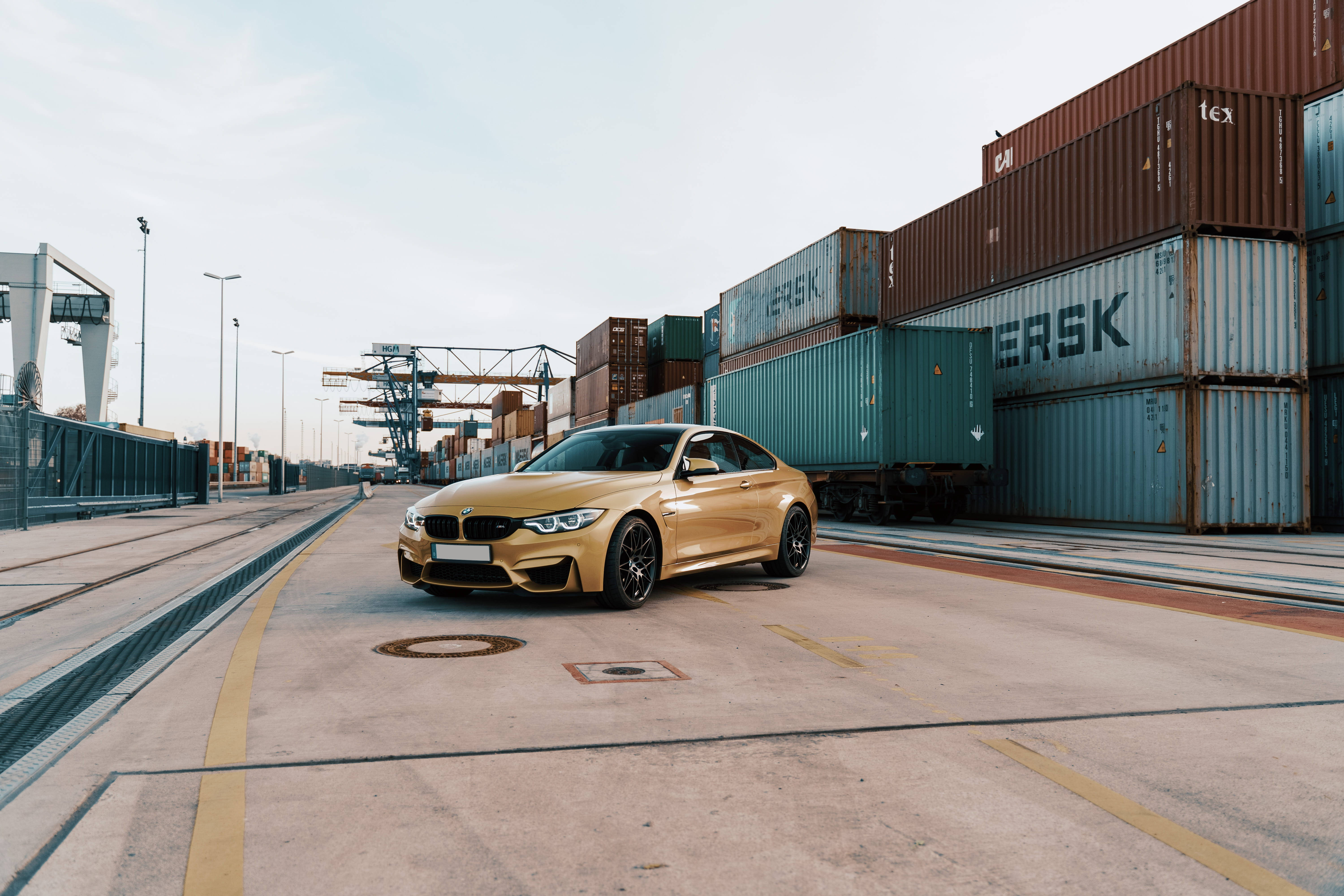 Sports Car 桌布 壁紙 — 一輛閃閃發光的金色BMW M4雙門跑車停泊在繁忙港口的水泥地上，周圍堆滿了貨運集裝箱。