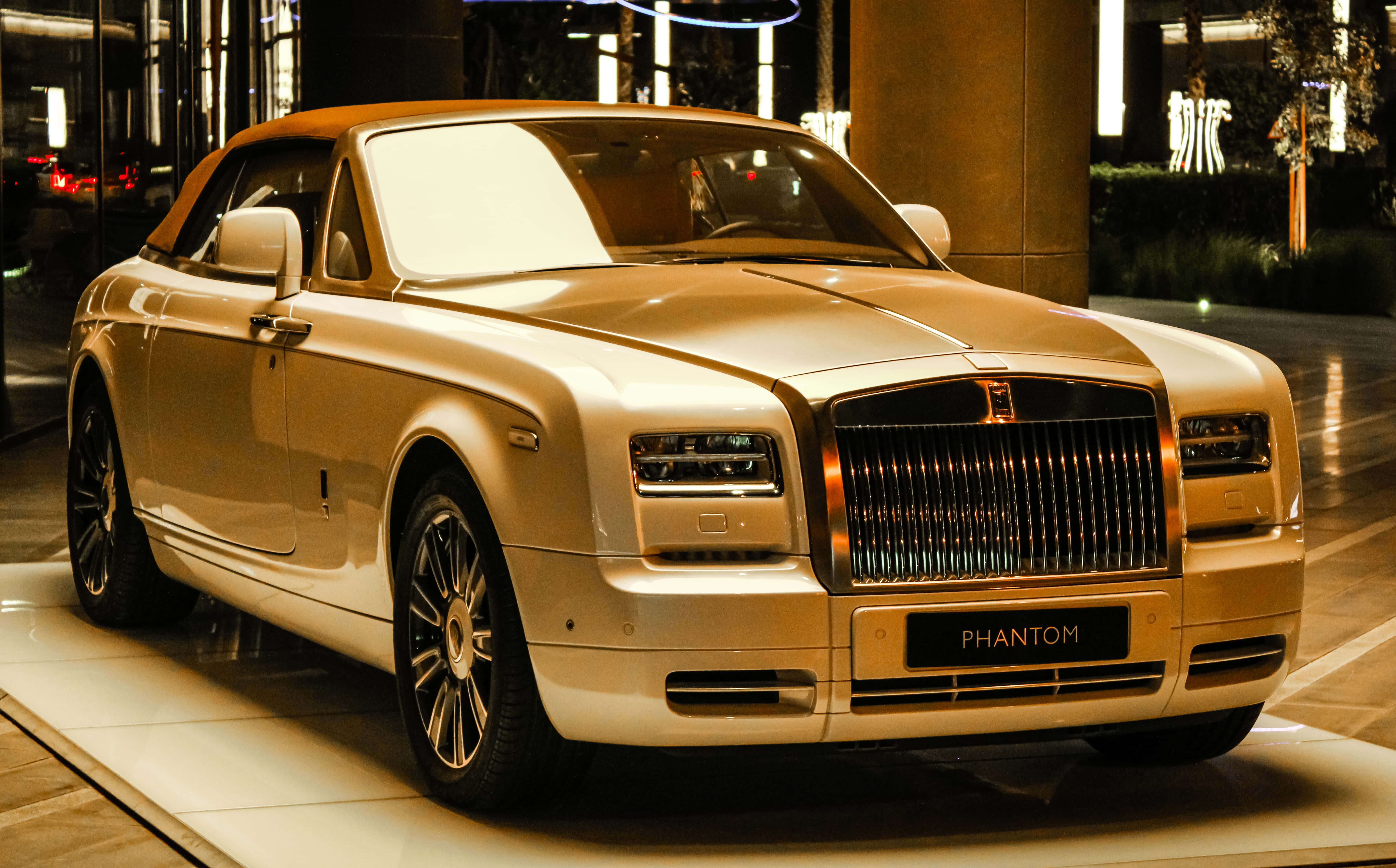Rolls Royce 桌布 壁紙 — 一輛閃閃發光的金色勞斯萊斯 Phantom 開篷車在溫暖的室內燈光下展示，背景有反射表面和現代建築。