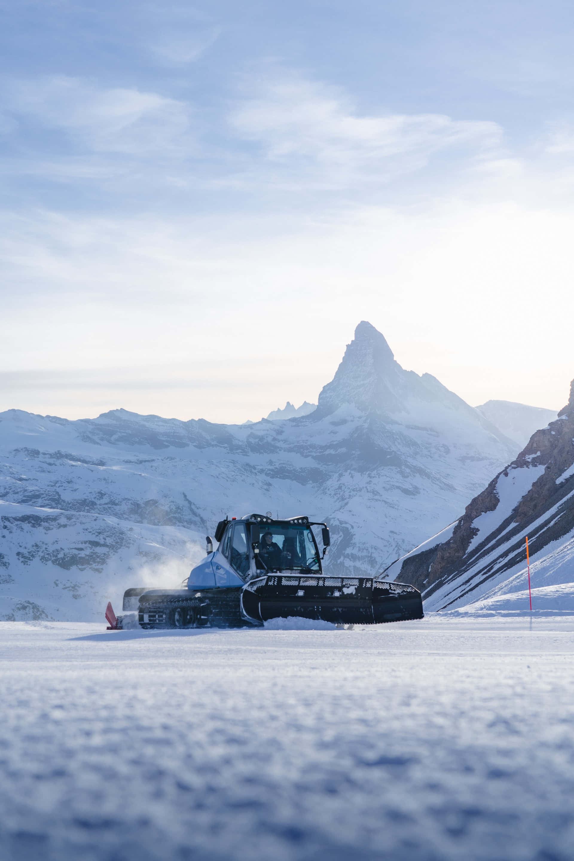 Snowplow 桌布 壁紙 — 一輛雪地車在純淨的雪中犁行，標誌性的馬特洪峰在柔和的藍天下雄偉地聳立在背景中。