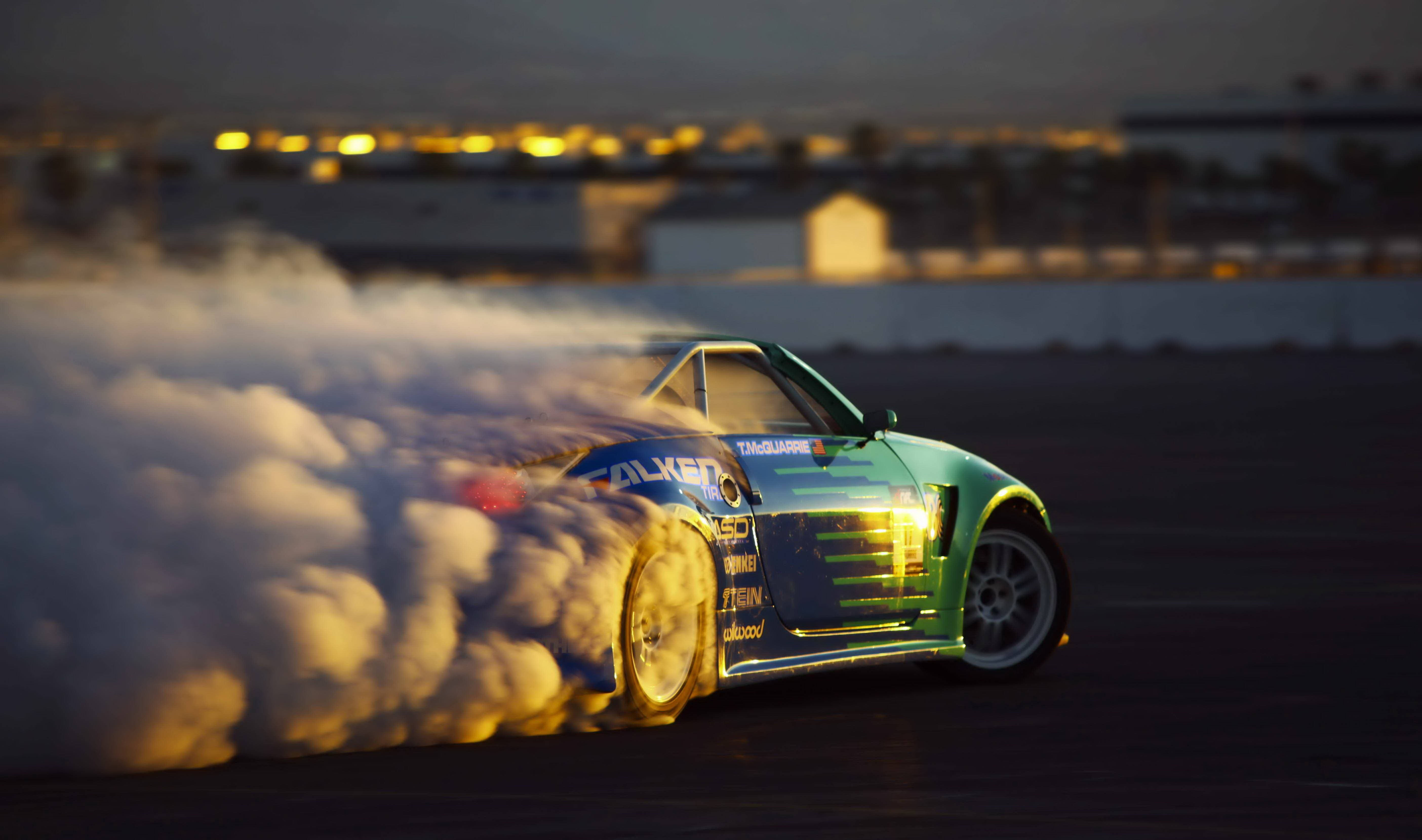 Sports Car 桌布 壁紙 — 一輛青綠色飄移車在暮色中於黑暗賽道上滑行，揚起濃煙，遠處城市燈光閃爍。