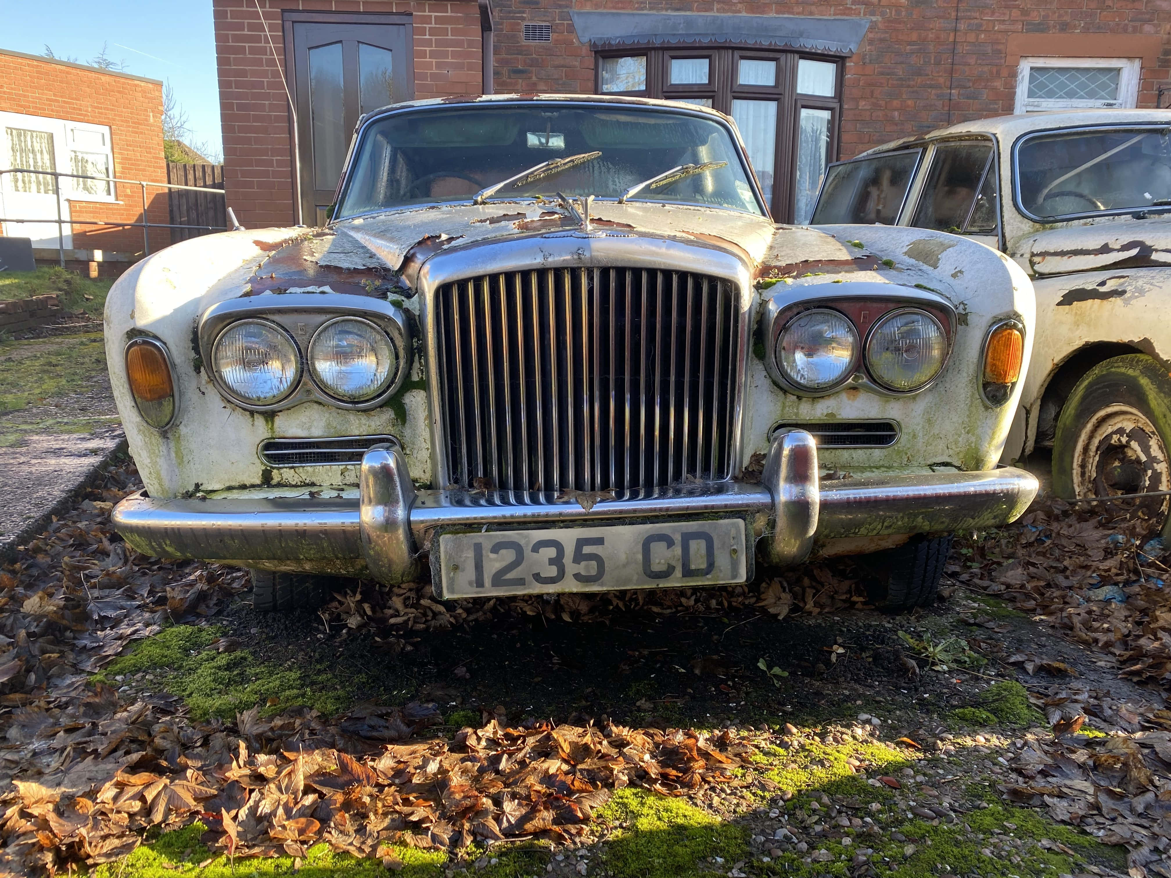 Bentley T1 桌布 壁紙 — 一輛風化的復古賓利，車身生鏽，地面長滿青苔，被遺棄在磚房旁邊一個落葉滿地的院子裡。