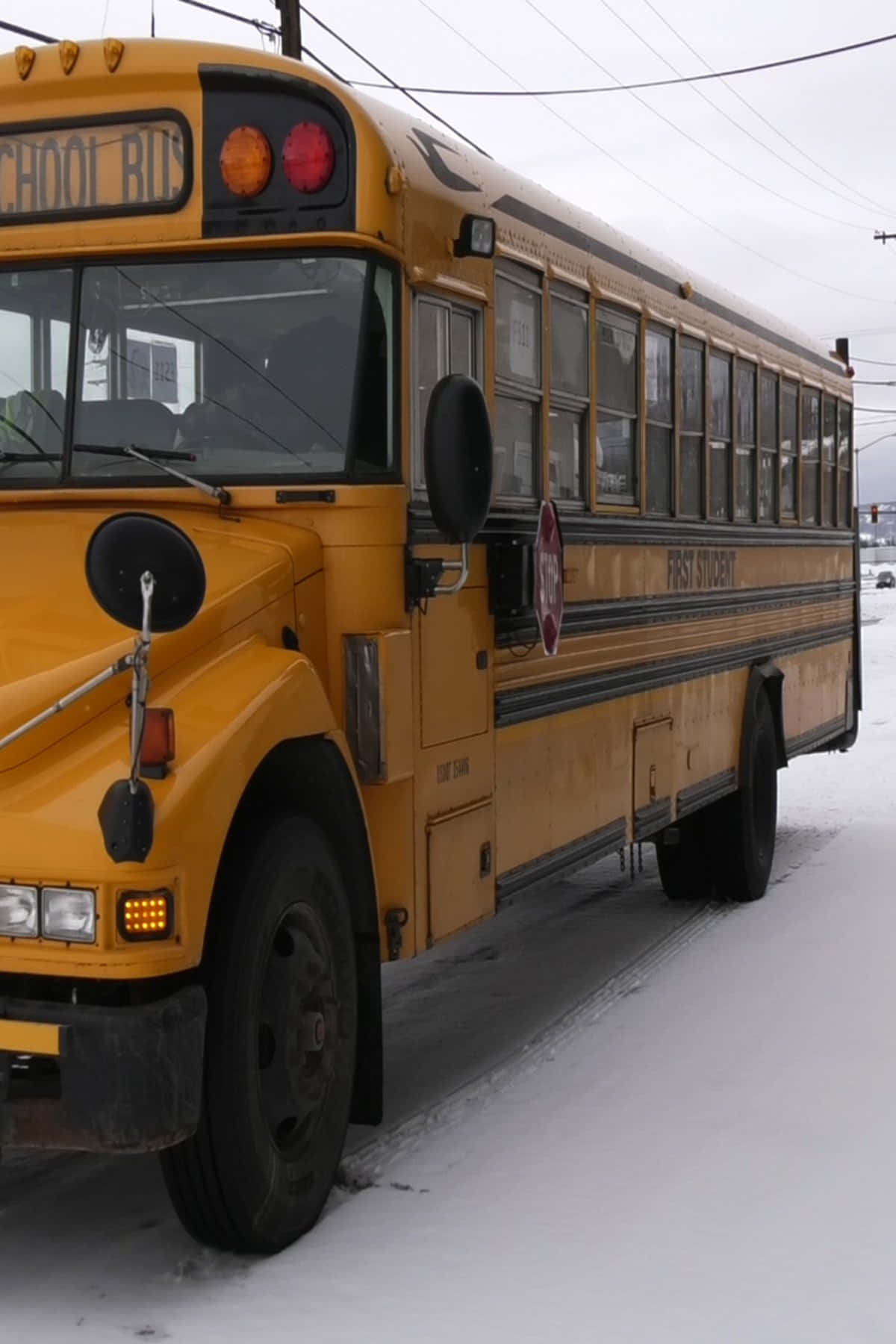 Yellow School Bus 桌布 壁紙 — 一輛風化的黃色校巴停泊在積雪的停車場，天空陰沉，頭頂有電線。
