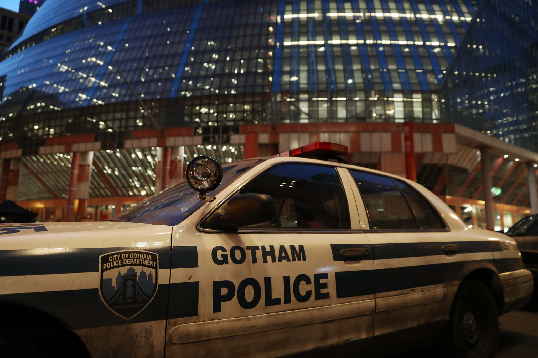 Gotham City Police Department 桌布 壁紙 — 一輛飽經風霜的葛咸城警車在夜晚停泊在發光的未來主義摩天大樓前。