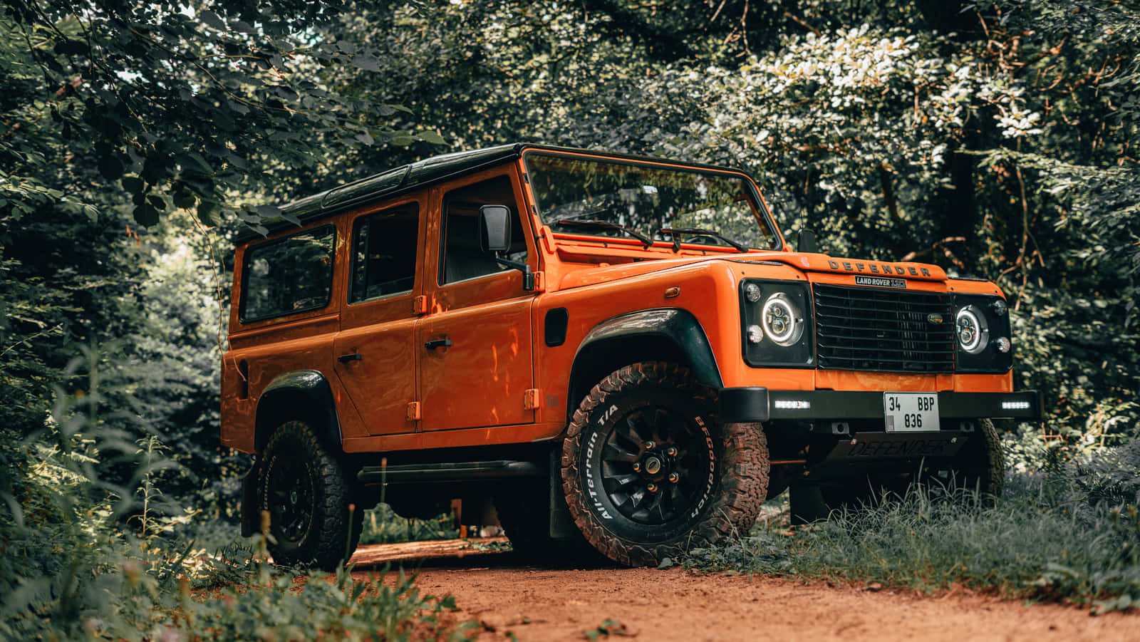 Land Rover Defender 桌布 壁紙 — 一輛鮮橙色的 Land Rover Defender 停泊在泥濘小徑上，四周被茂密的綠色森林環繞。
