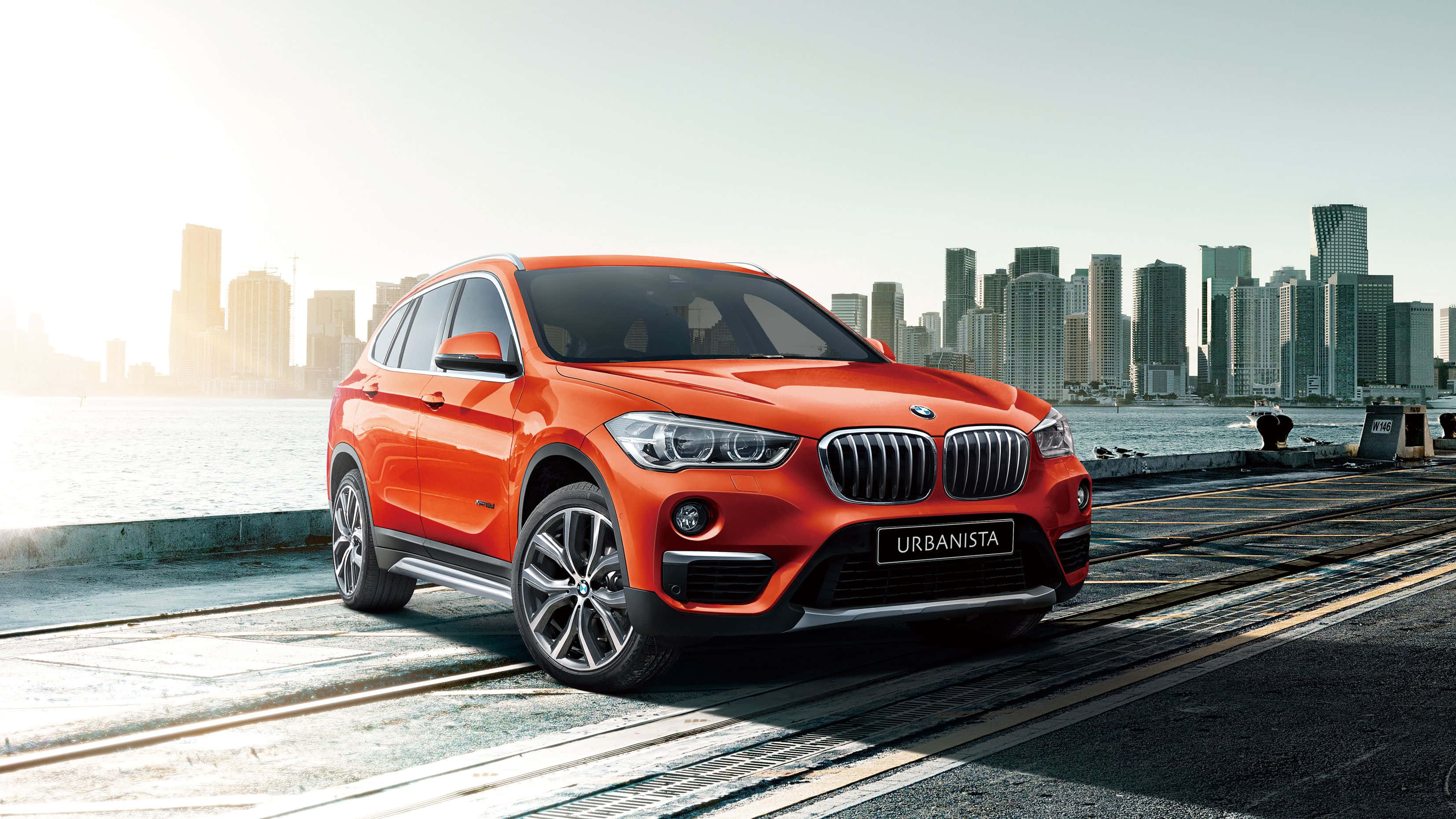 Bmw X1 桌布 壁紙 — 一輛鮮橙色的BMW X1 Urbanista SUV停在海濱長廊上，背景是現代化的城市天際線。