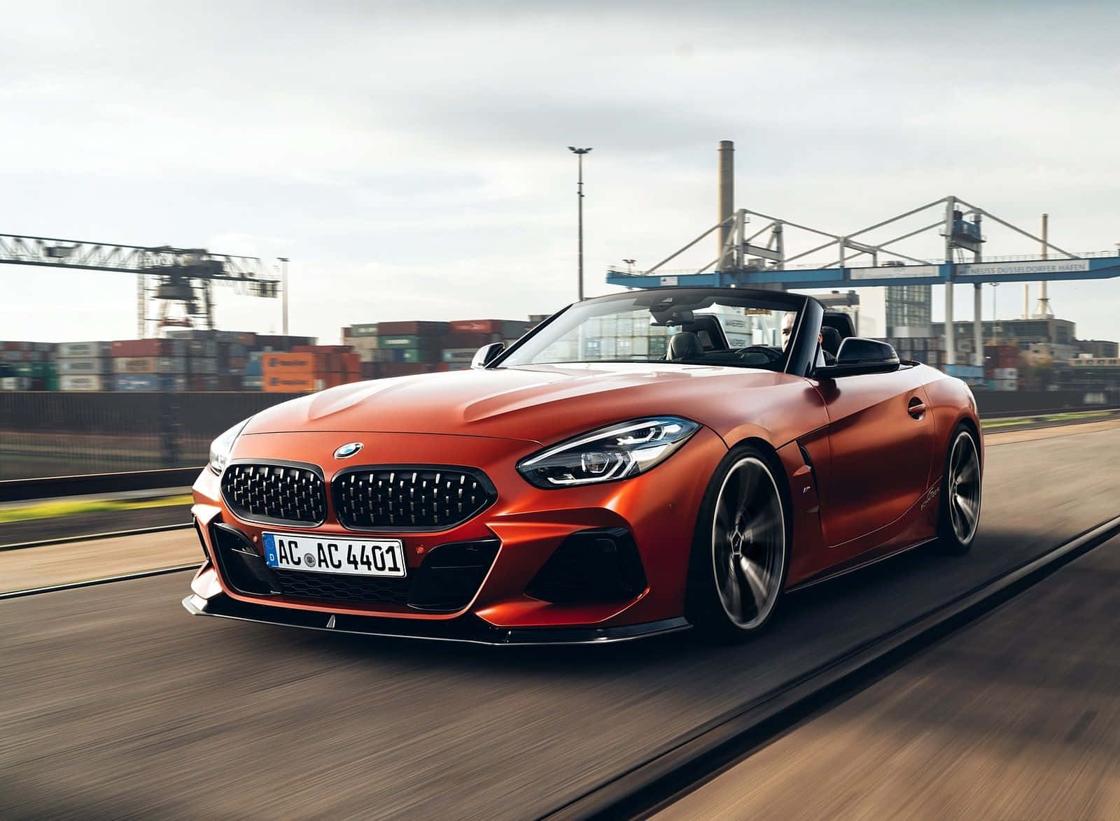 Bmw Z4 桌布 壁紙 — 一輛鮮橙色的BMW Z4開篷跑車在高速公路上飛馳，背景模糊中可見工業起重機和貨運集裝箱。