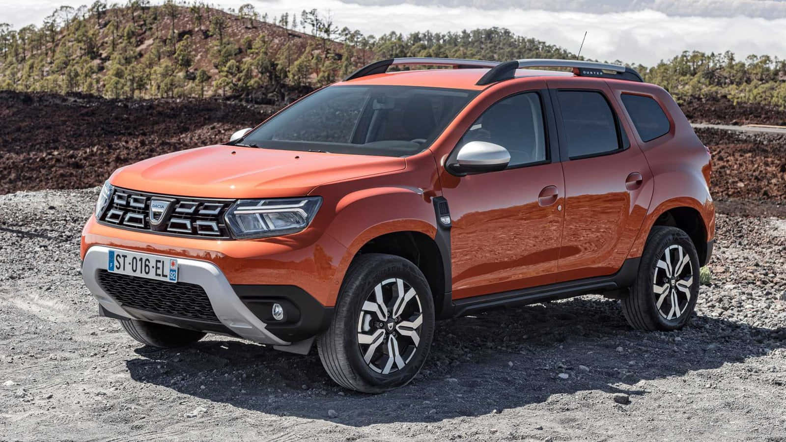 Dacia 桌布 壁紙 — 一輛鮮橙色的Dacia Duster SUV停泊在碎石上，背景是陽光明媚下的森林山丘。