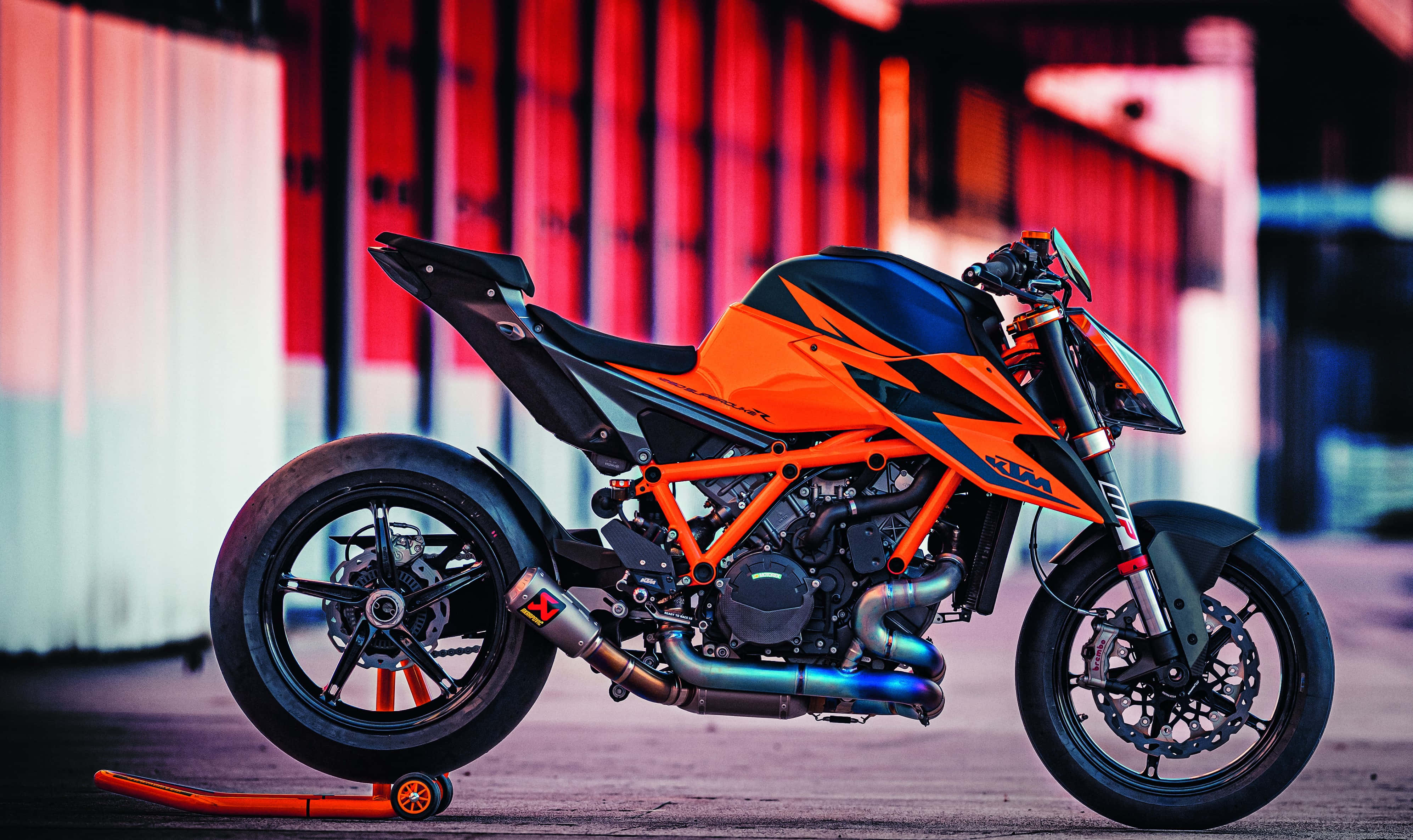 Ktm Bike 桌布 壁紙 — 一輛鮮橙色的KTM電單車停在支架上，背景是模糊的紅白工業場景。