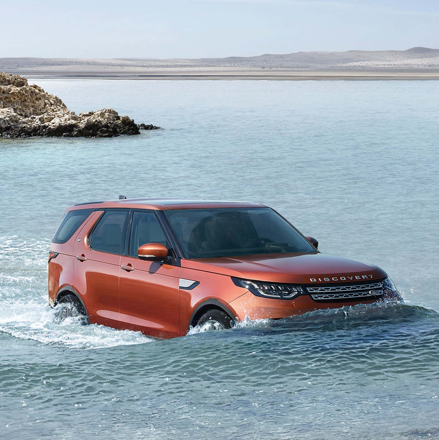 Land Rover Discovery 桌布 壁紙 — 一輛鮮橙色的Land Rover Discovery在晴朗天空下，靠近岩石海岸行駛於淺藍綠色水中。