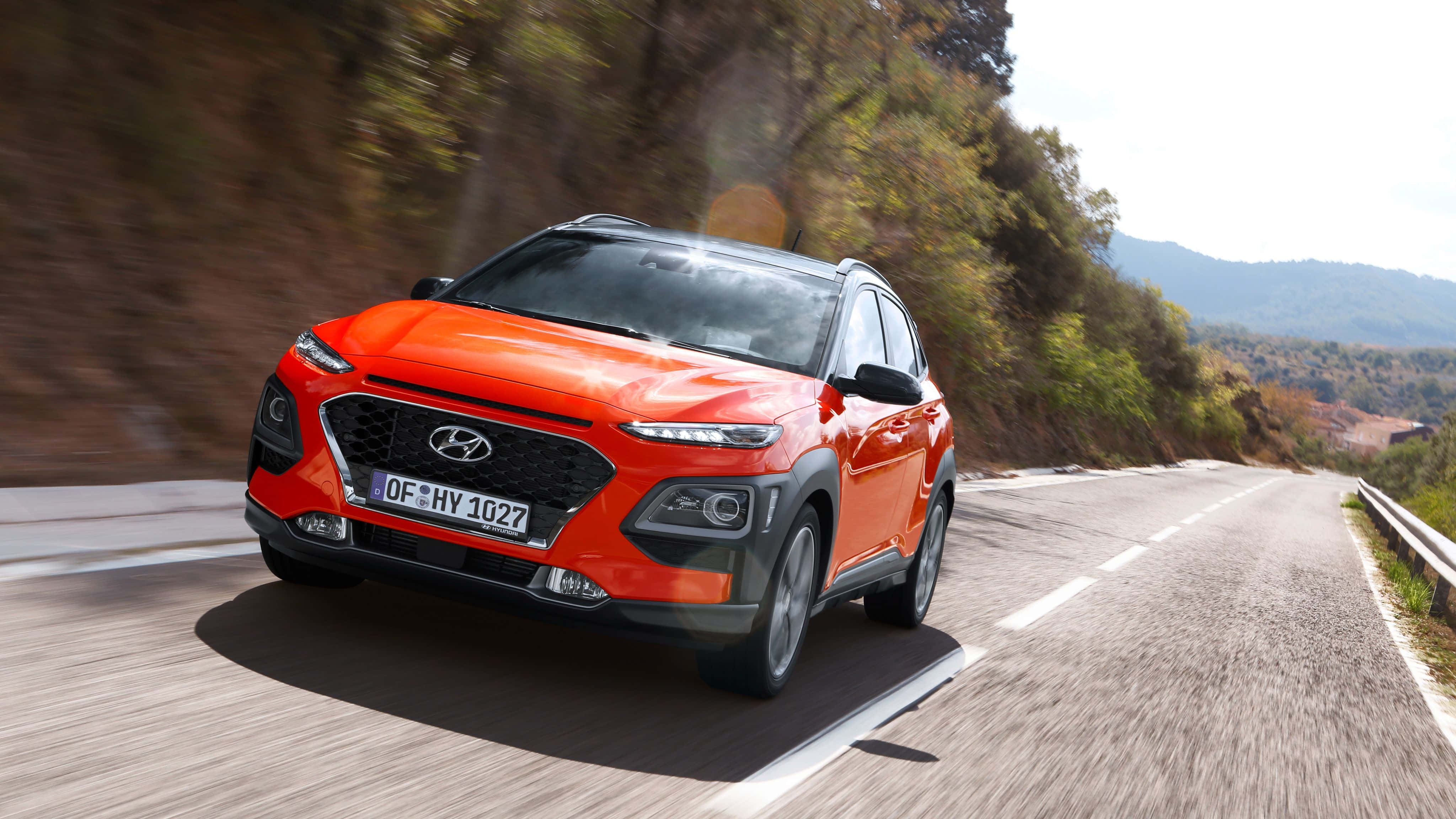 Hyundai Kona 桌布 壁紙 — 一輛鮮橙色的現代Kona SUV在明亮的日光下，沿著蜿蜒的山路飛馳，背景是模糊的綠樹和遠山。