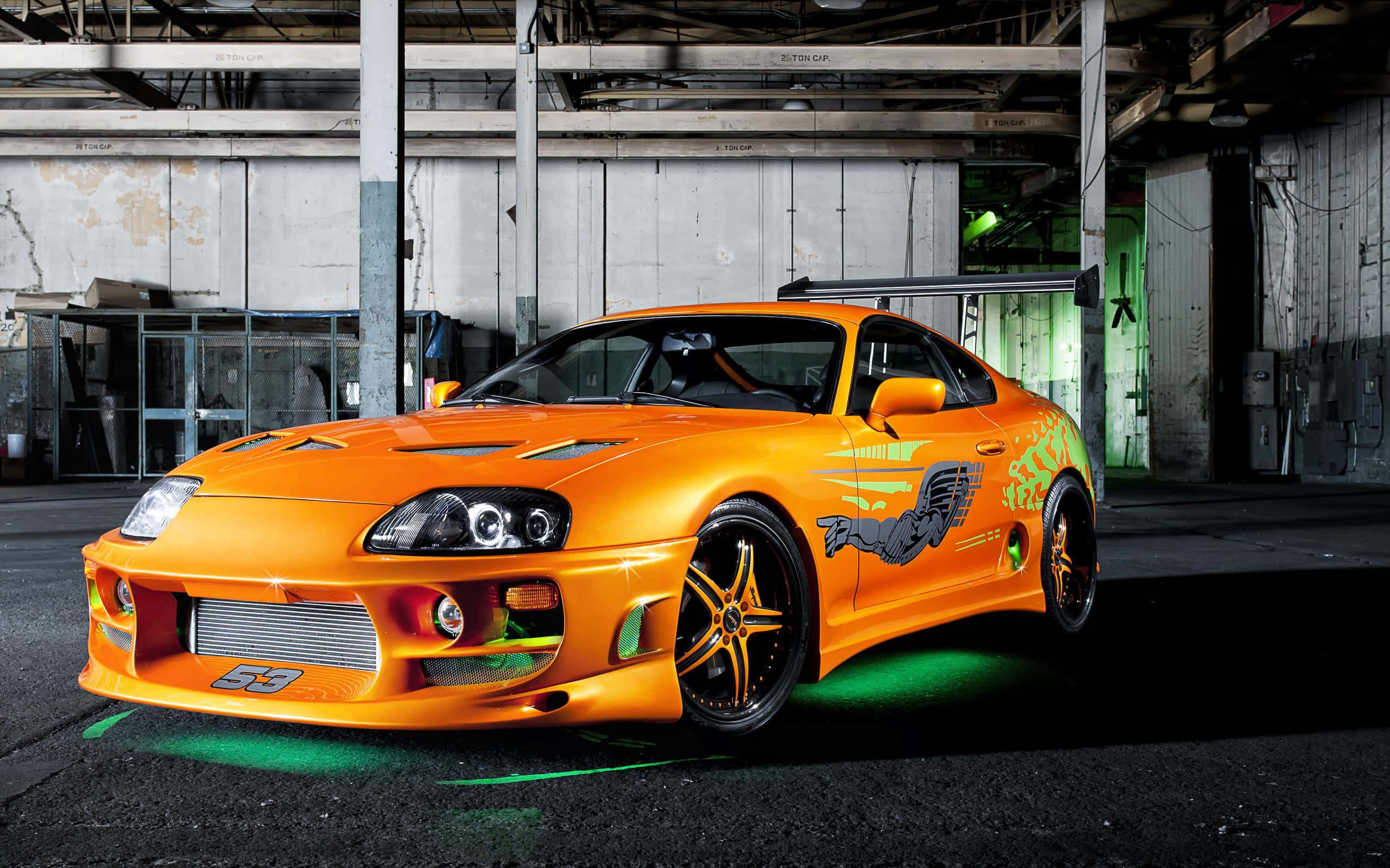 極致力量——非凡改裝跑車 壁紙 — 一輛鮮橙色豐田Supra，配有綠色底盤燈和黑色輪圈，停放在骯髒的工業倉庫中。