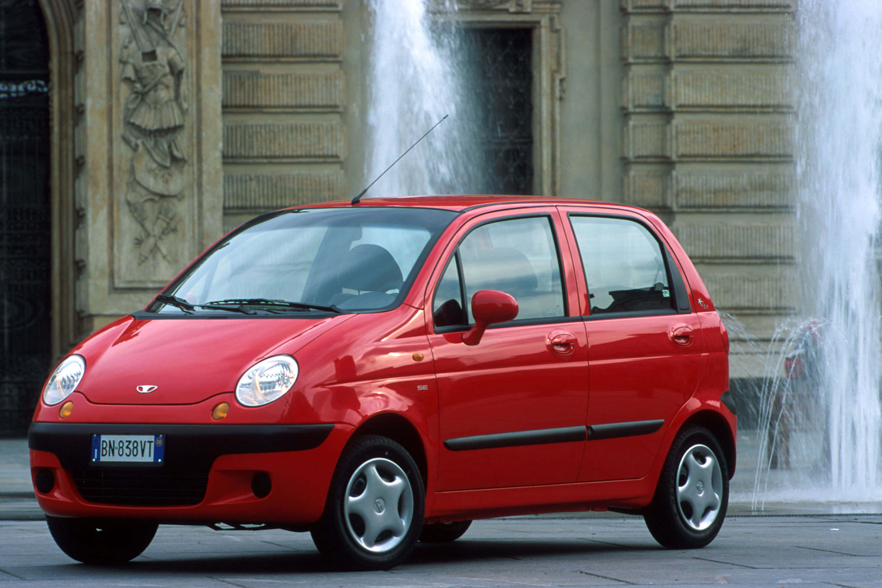 Daewoo 桌布 壁紙 — 一輛鮮紅色嘅小型房車停喺宏偉建築物前面，後面有活躍嘅噴泉喺度噴水。