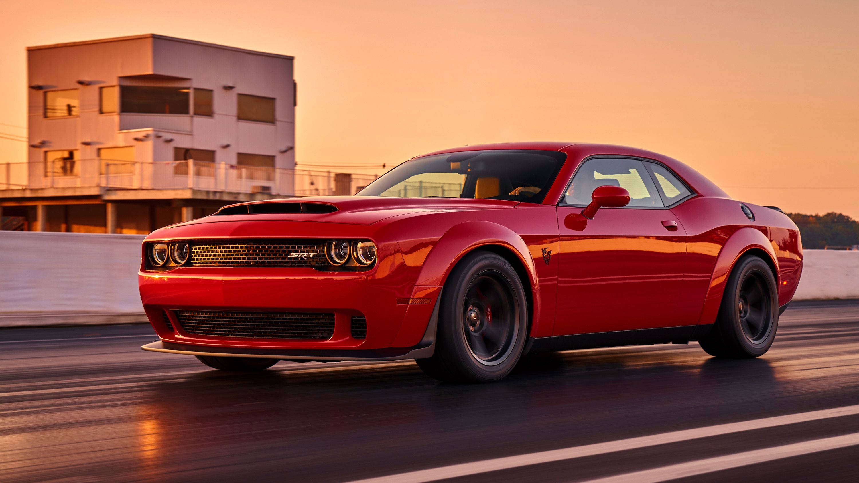 Dodge Challenger SRT Demon 10 壁紙 — 一輛鮮紅色的 Dodge Challenger 在日落時分沿著賽道飛馳，背景是一座現代建築。