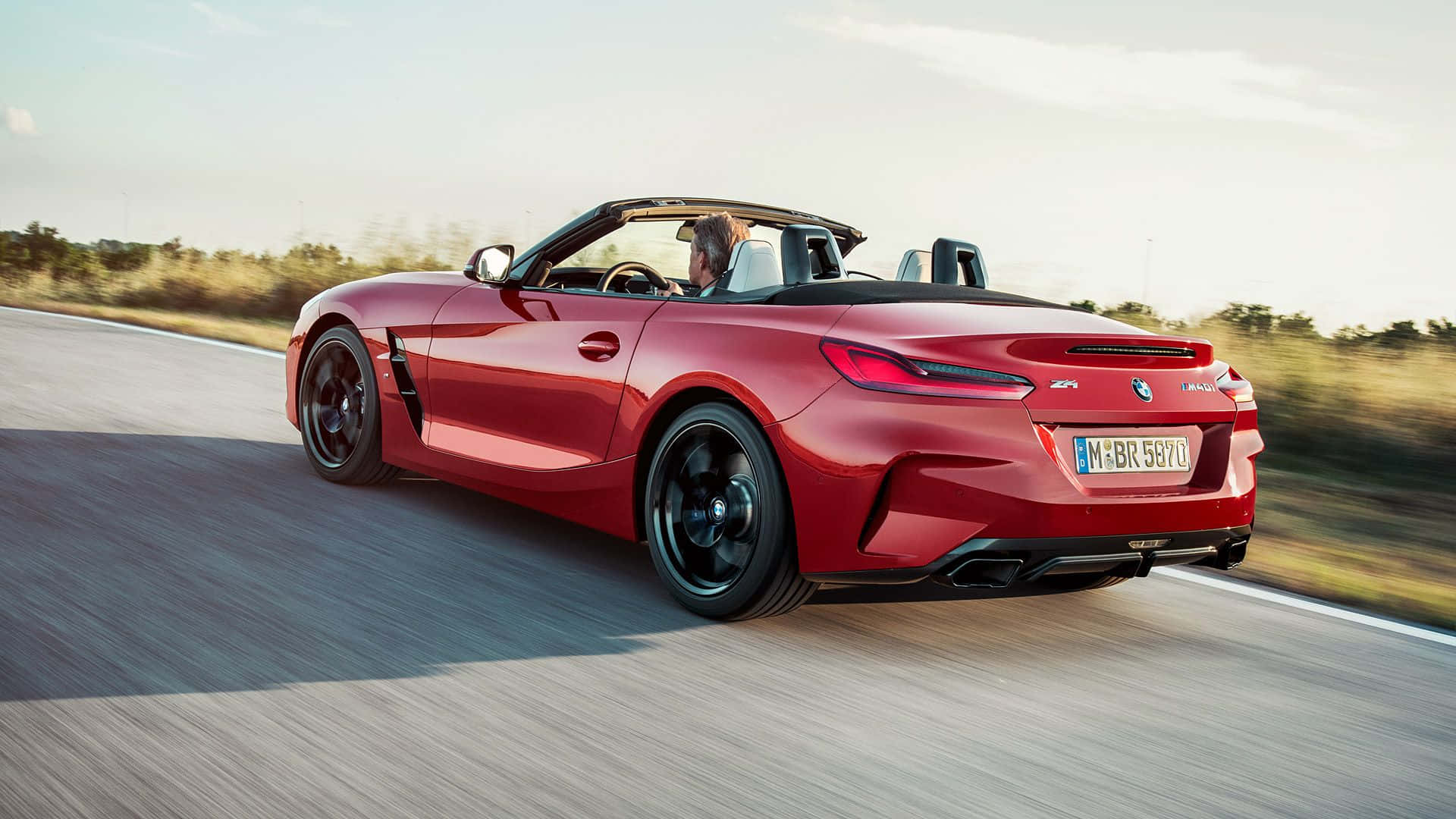Bmw Z4 桌布 壁紙 — 一輛鮮紅色的BMW Z4開篷跑車在陽光明媚的道路上行駛，背景有模糊的動態和金色的樹葉。